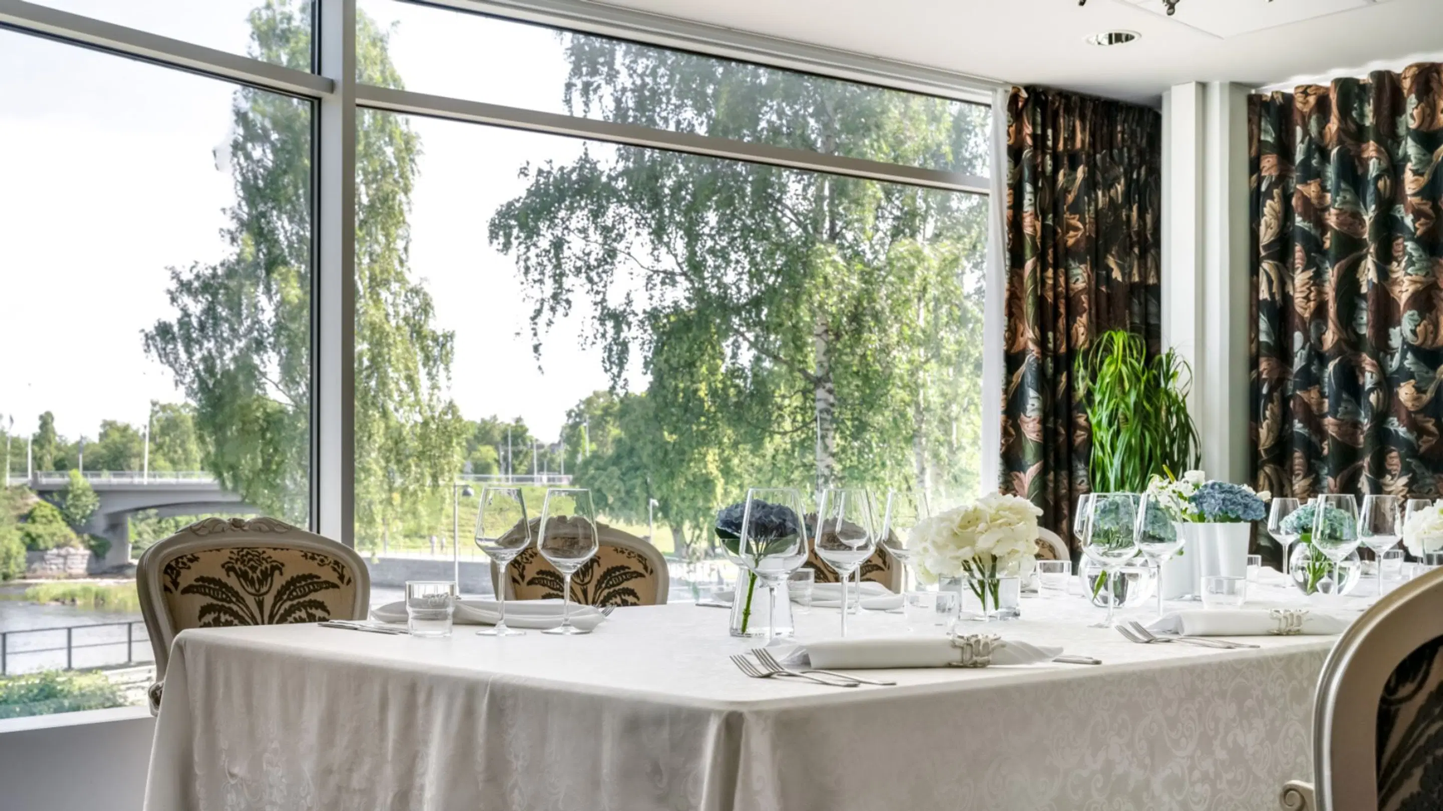 Elegant restaurant table with white tablecloth and crystal glasses. Large windows overlook lush green parkland and a bridge.