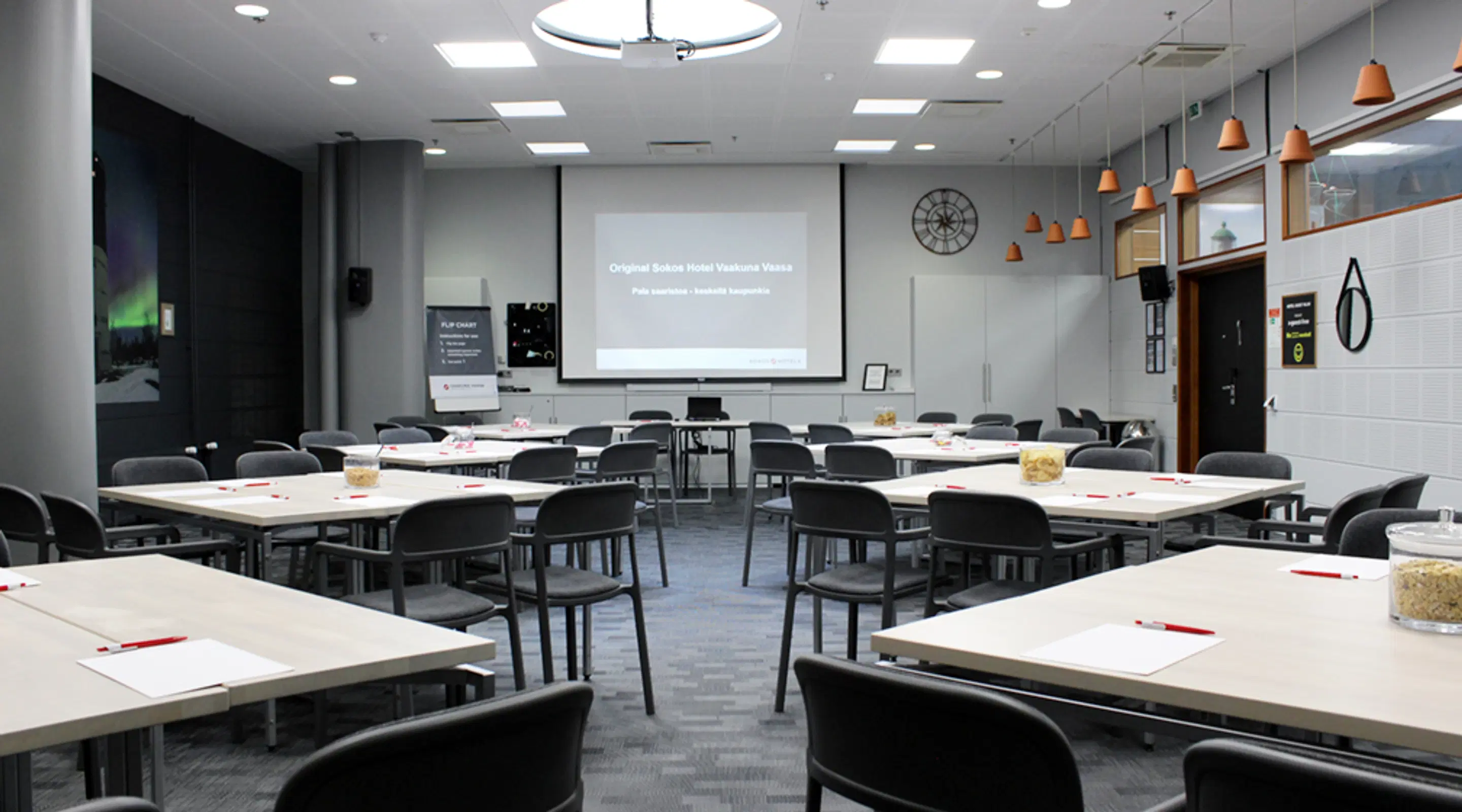 Modern conference room with white tables, black chairs and a presentation screen. The room is well-lit with ceiling lights and orange pendant lamps.