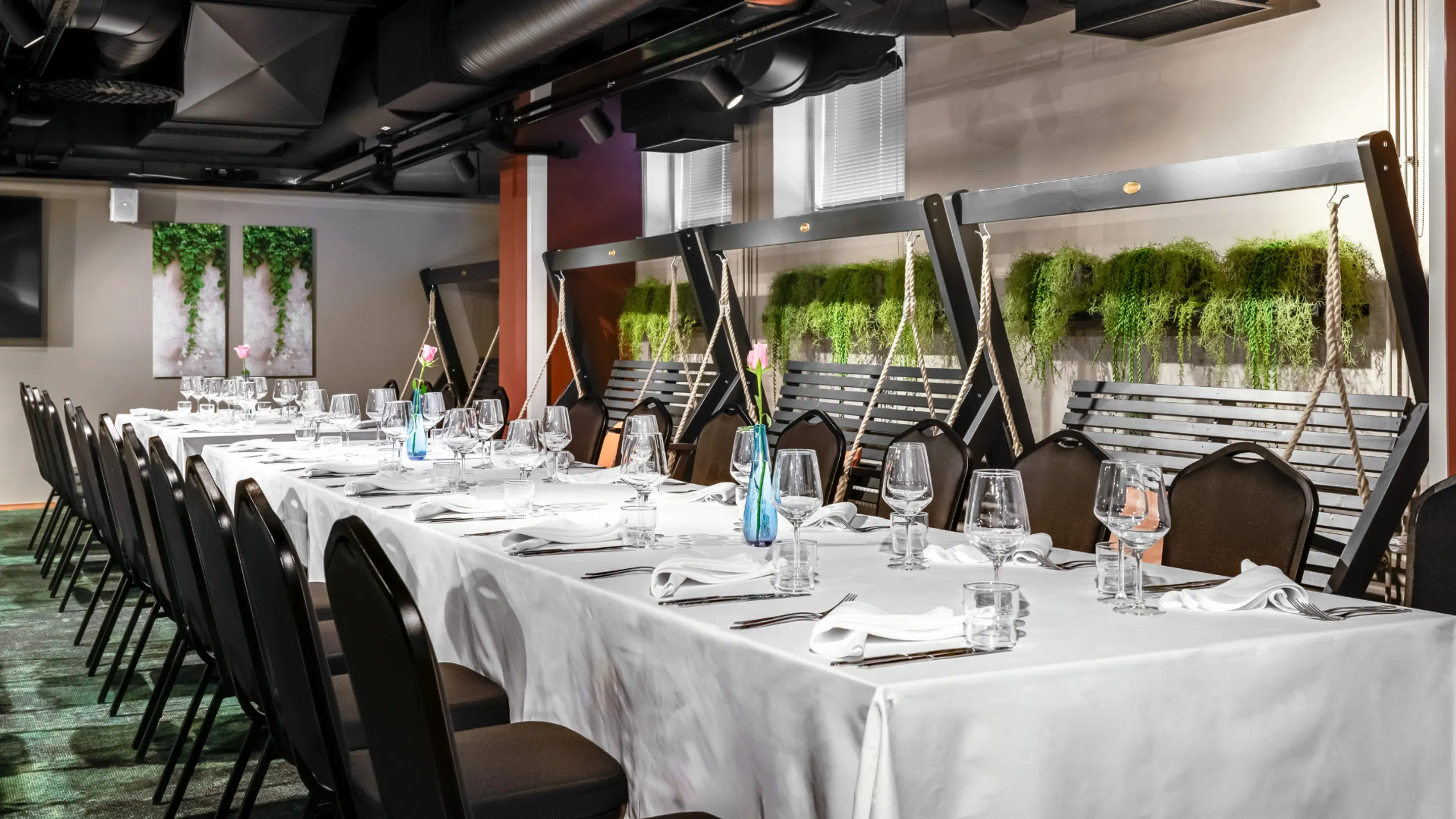 Modern dining room with a long table covered in white cloth and decorative swings along the wall. The space is adorned with hanging greenery.