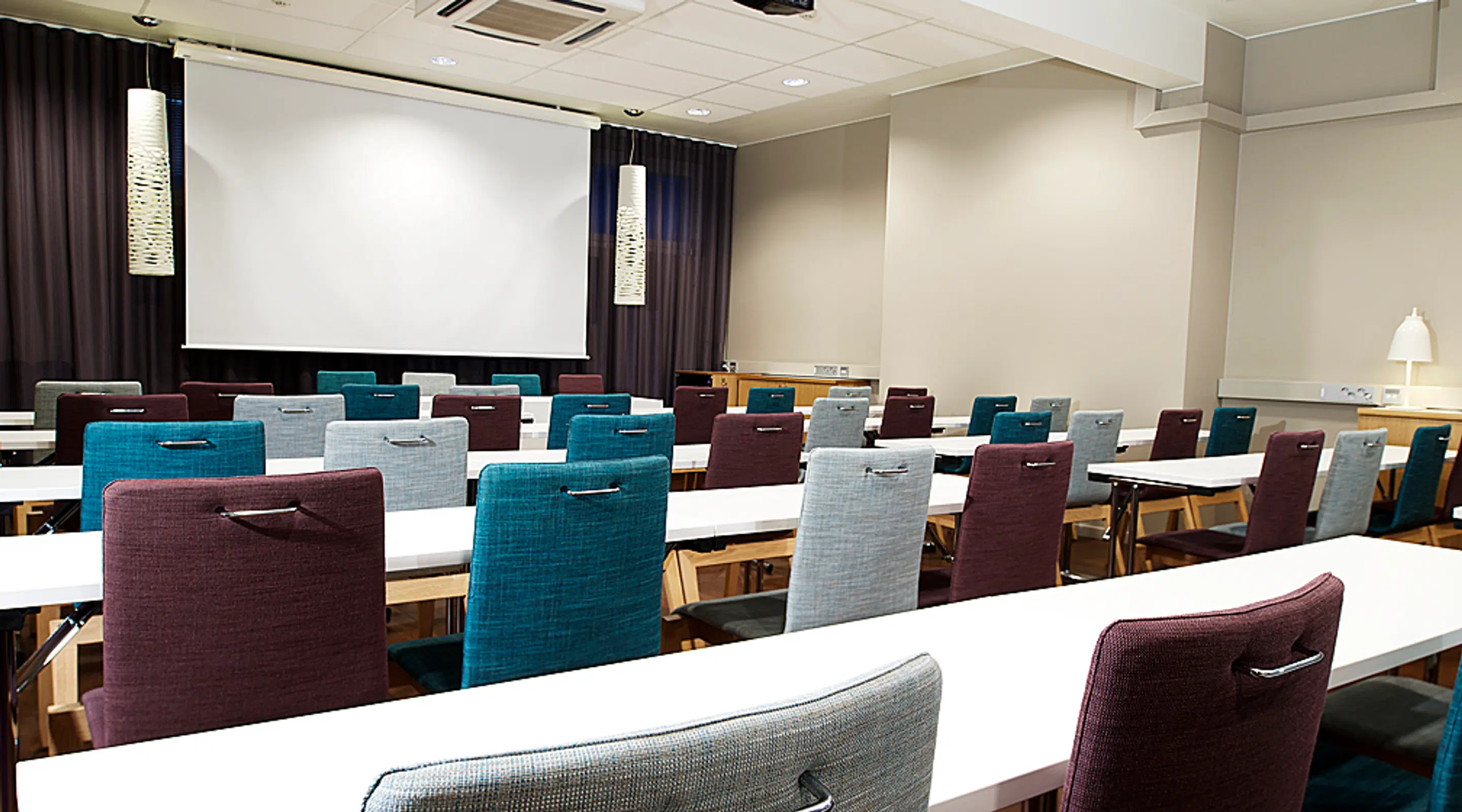 Conference room with white tables and colorful chairs in turquoise, gray, and burgundy. A projection screen and dark curtains at the front.