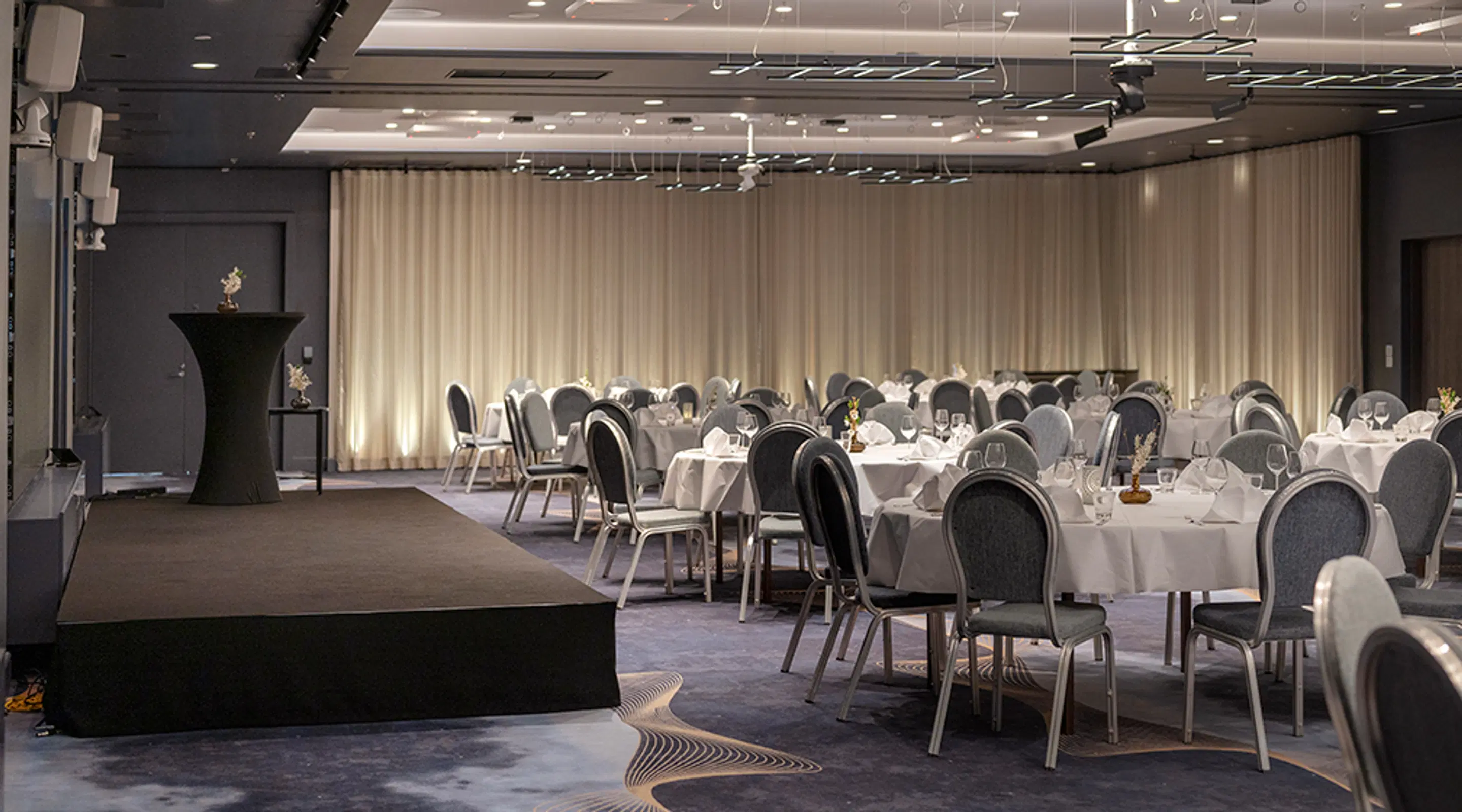 Banquet hall with round set tables and upholstered chairs. The hall has a low stage and a tall cocktail table. The floor has a patterned carpet. Lighting is soft and warm.