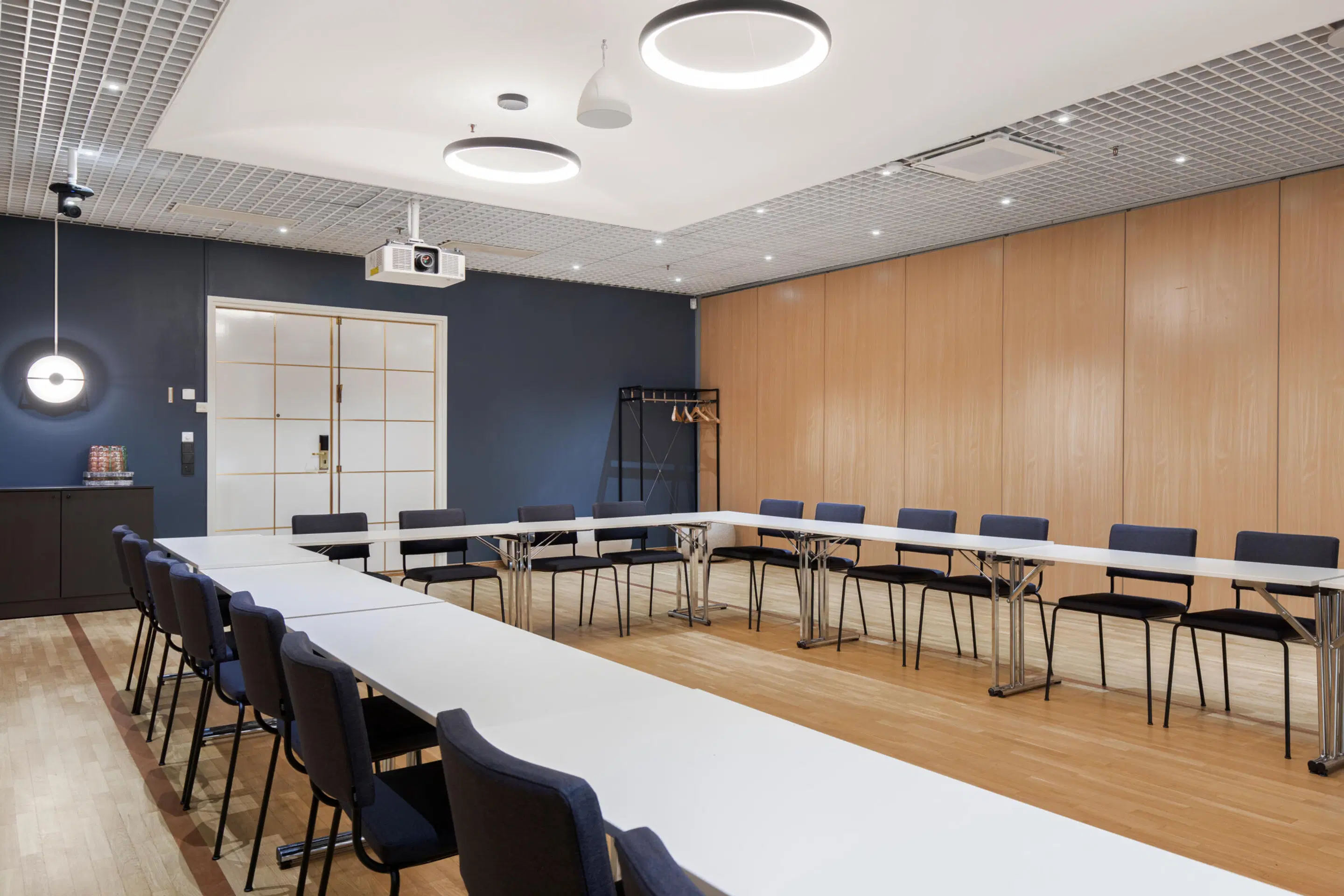 Modern meeting room with U-shaped table arrangement, blue wall and wood paneling. Space features stylish lighting and wooden flooring.
