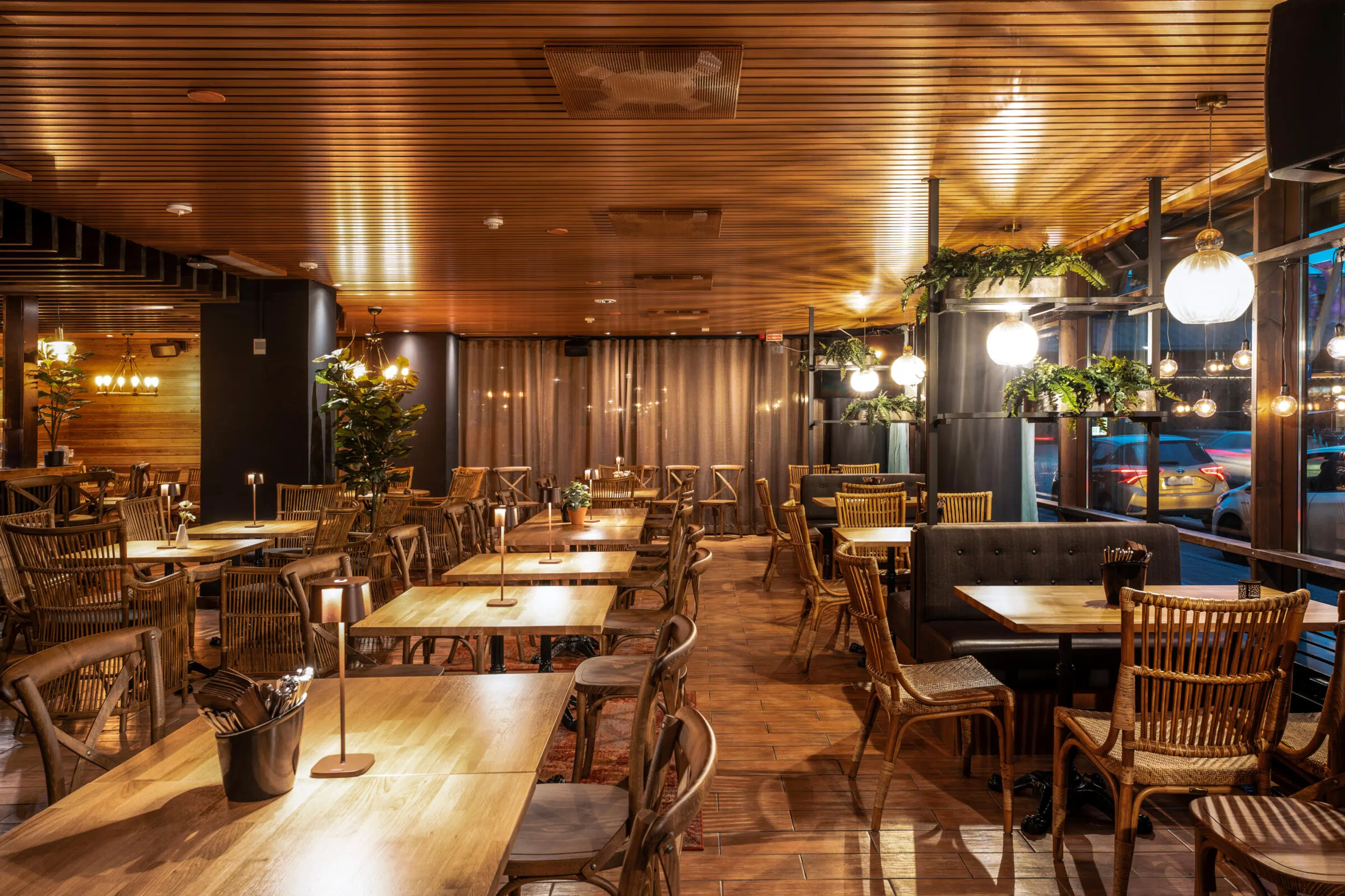 Restaurant interior with wooden ceiling, rattan chairs and wooden tables. Warm lighting, green plants and window facing the street.