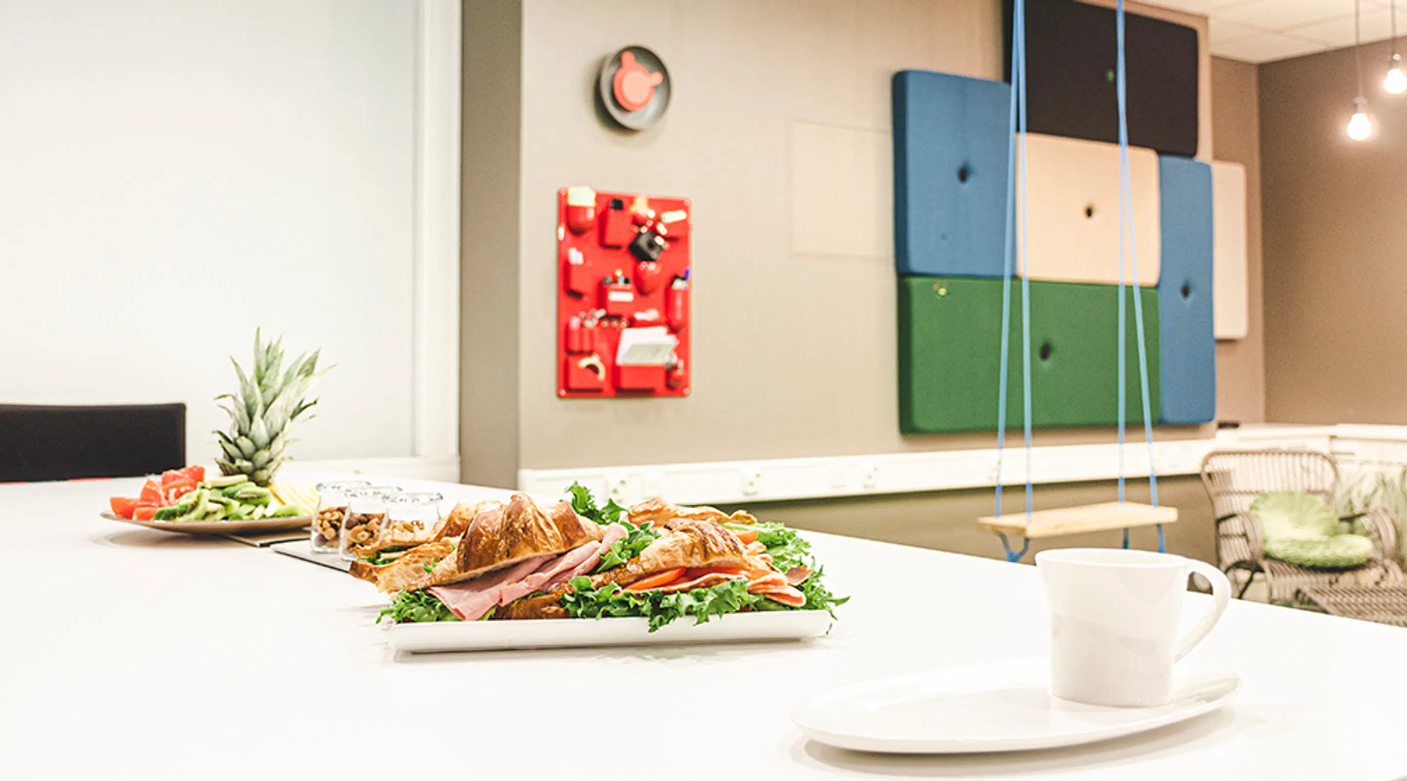 White table with a croissant sandwich, fruit platter and coffee cup. Colorful acoustic wall panels and a clock in the background.