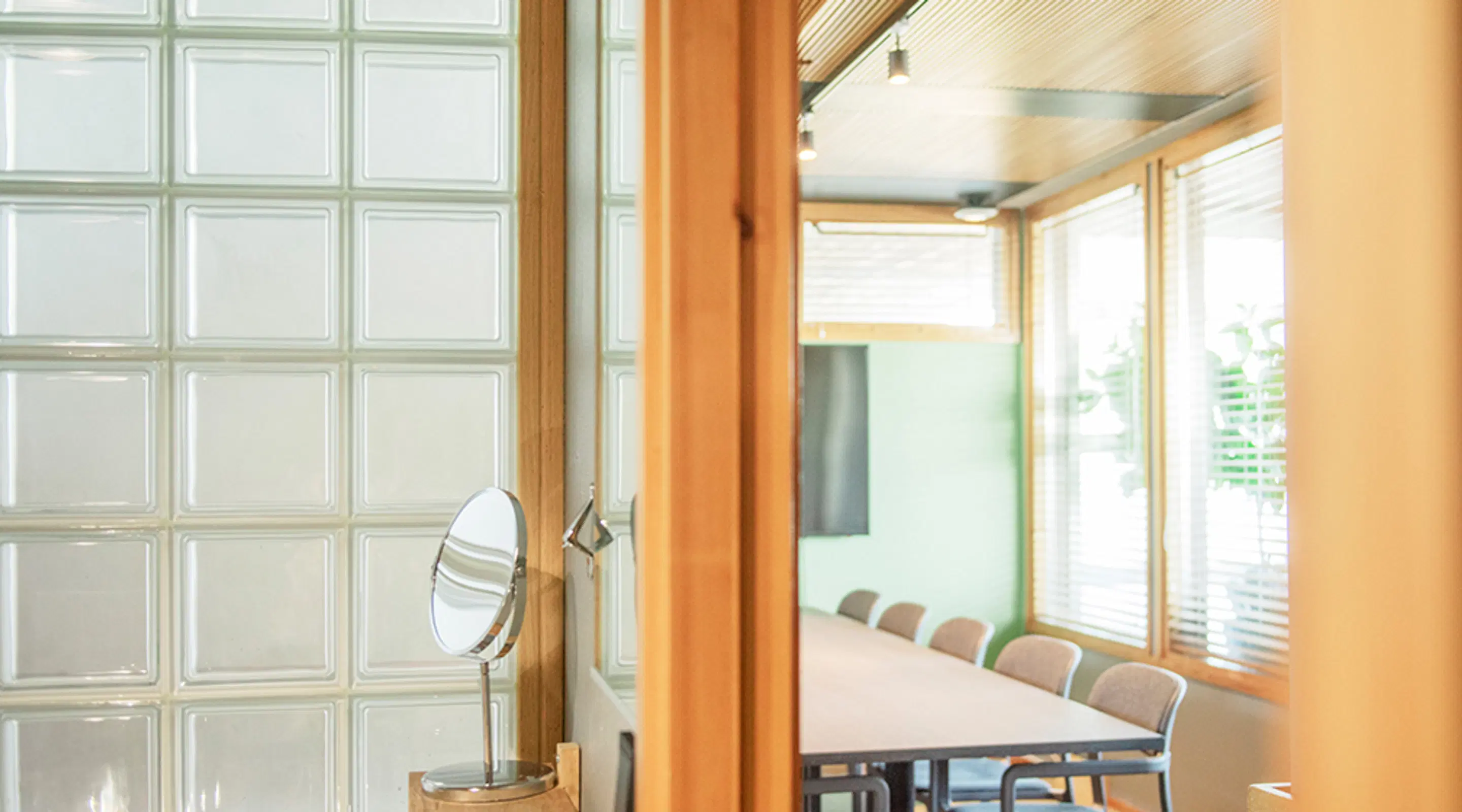 Meeting room viewed through a glass wall. Inside, a long table with several chairs and bright windows with blinds.