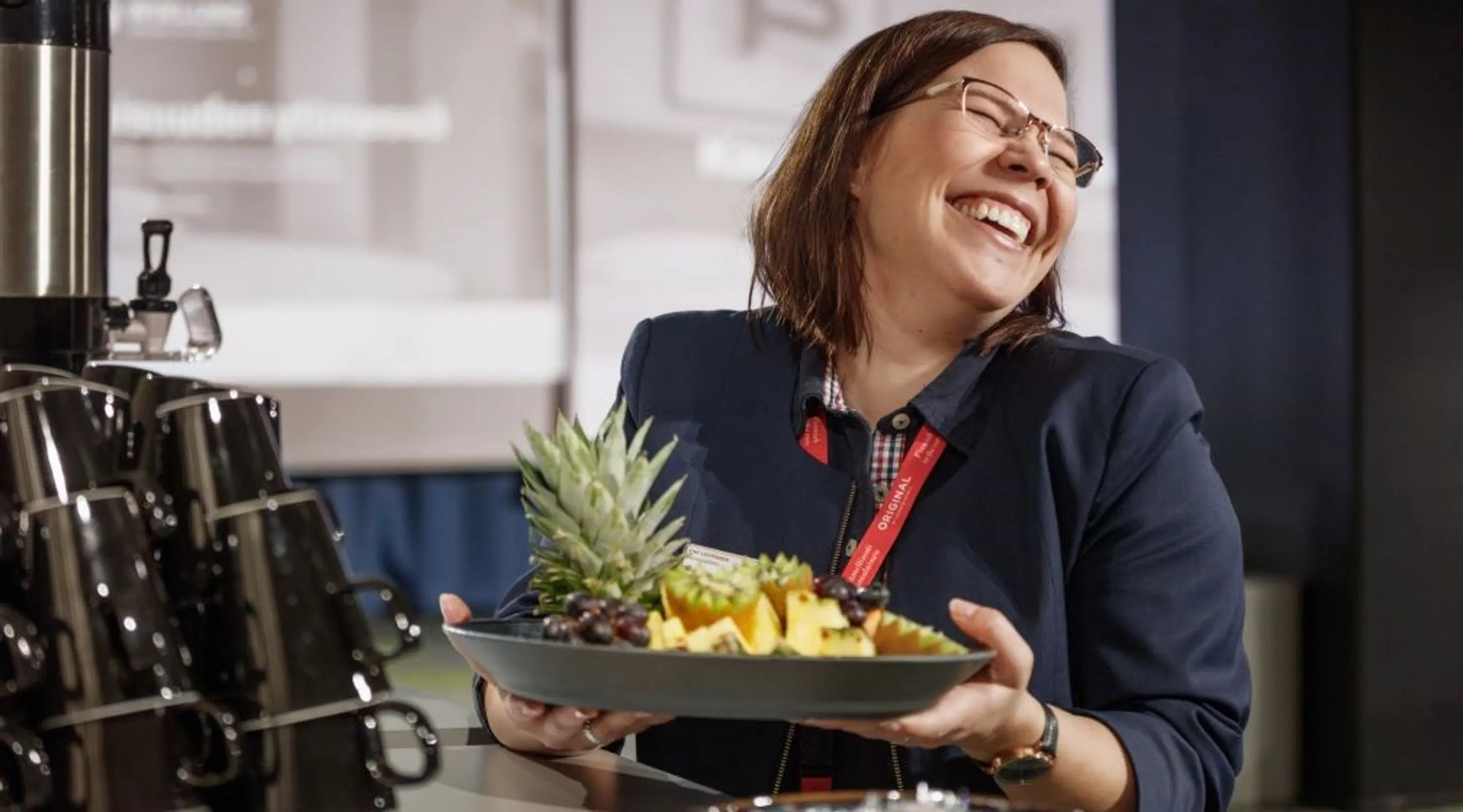 Person i blå kavaj håller en tallrik med ananas och annan frukt bredvid en kaffeautomat.