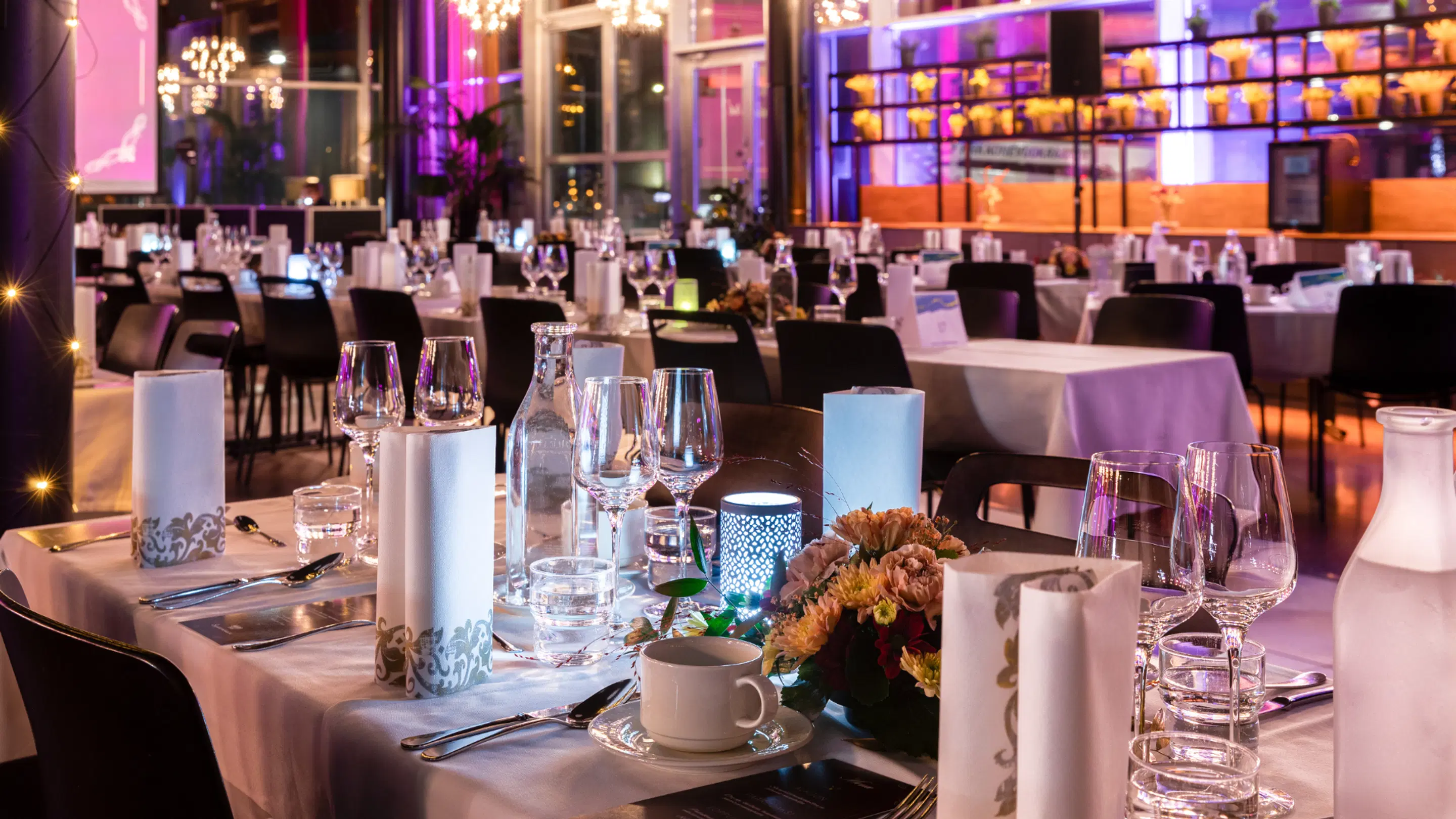 Elegantselt kaetud restoran valgete laudlinadega laudade, joogiklaaside ja lilleseadetega. Taustal roosa ja lilla valgustus.