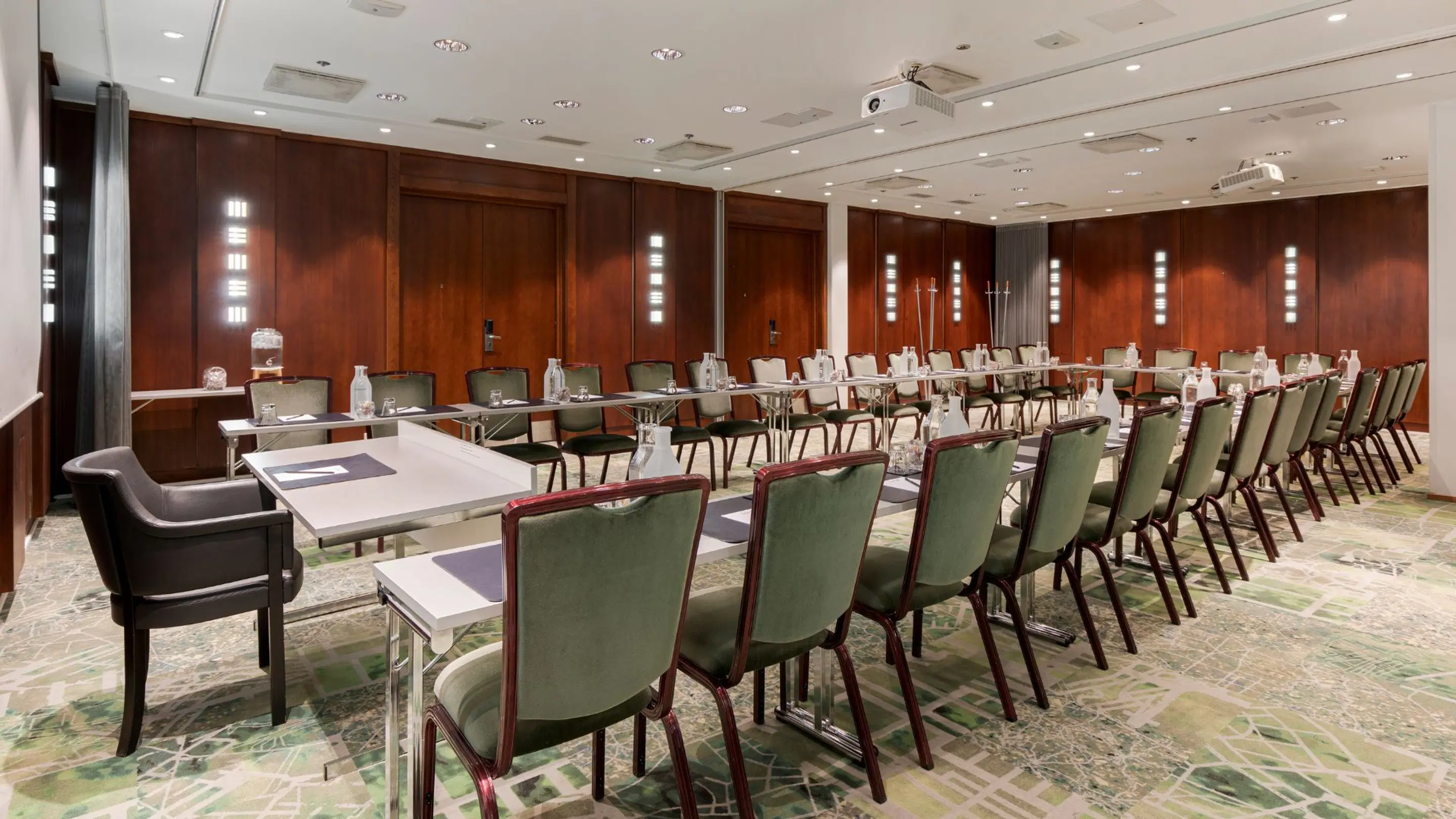 Conference room with green chairs and white tables arranged in U-shape. Room features wooden paneled walls and flat, marble-like flooring.