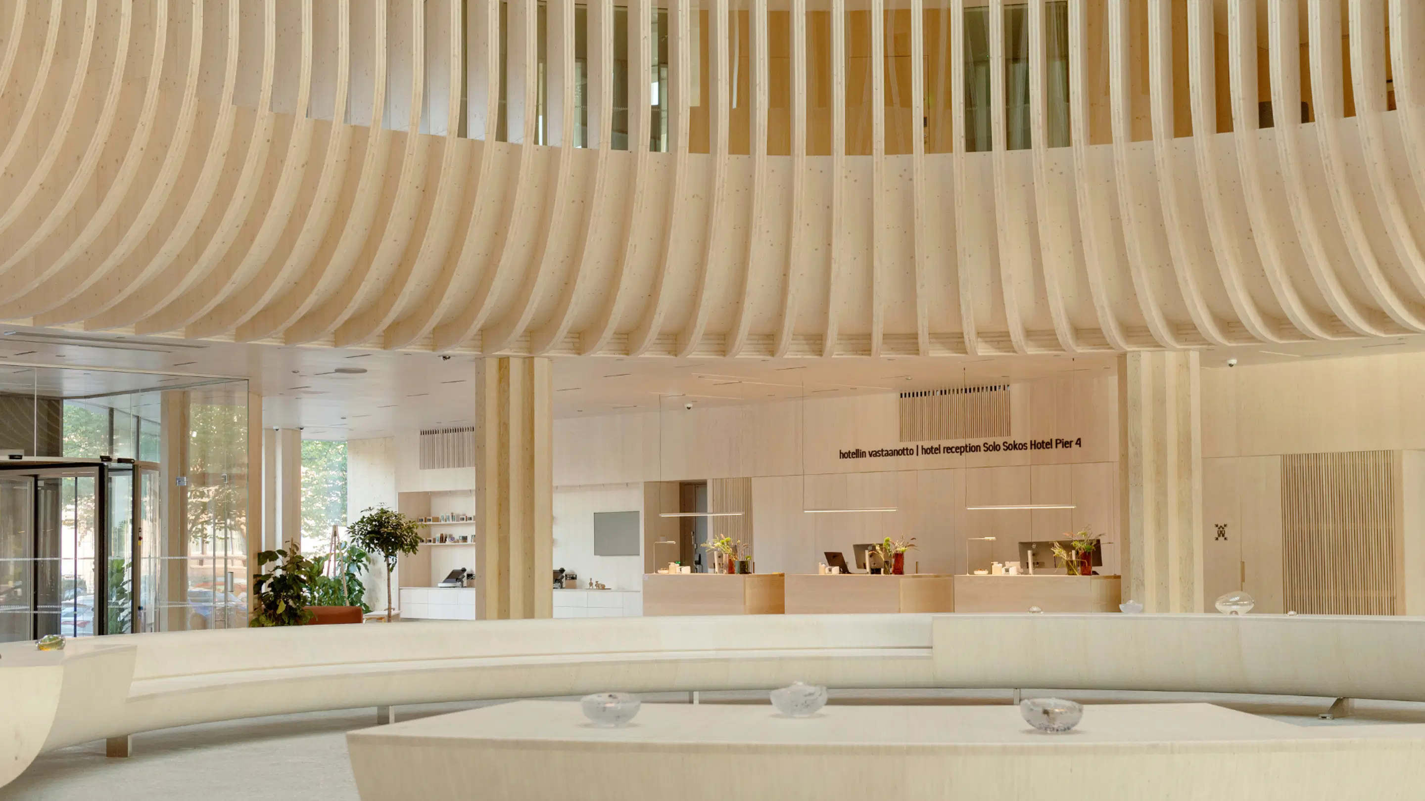 Modern hotel lobby featuring light wooden curved ceiling, reception desk and glass walls. Spacious area with natural light and indoor plants.
