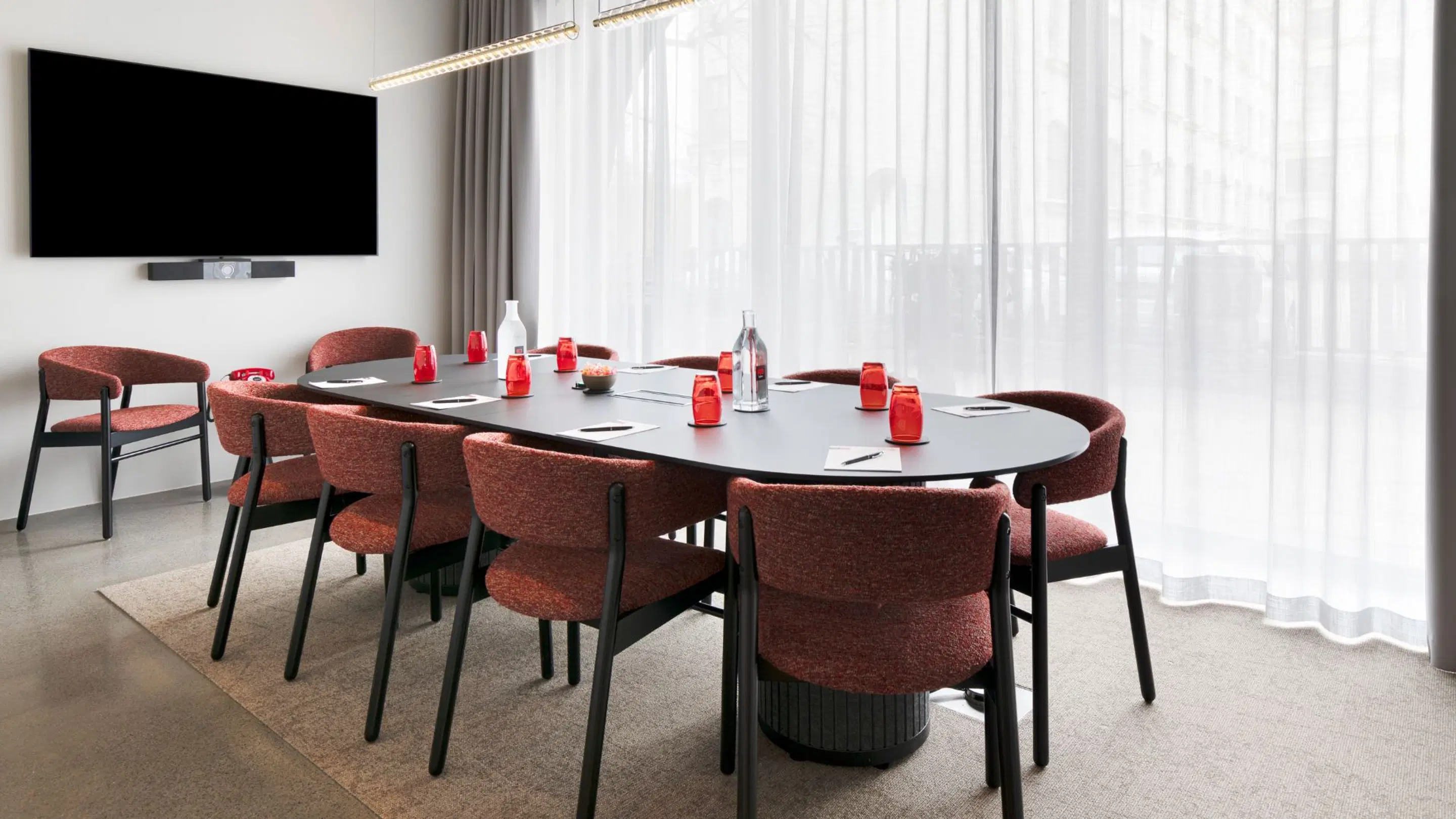 Modern meeting room with oval black table and reddish-brown chairs. Wall-mounted flat screen, large windows, and level carpeted floor.