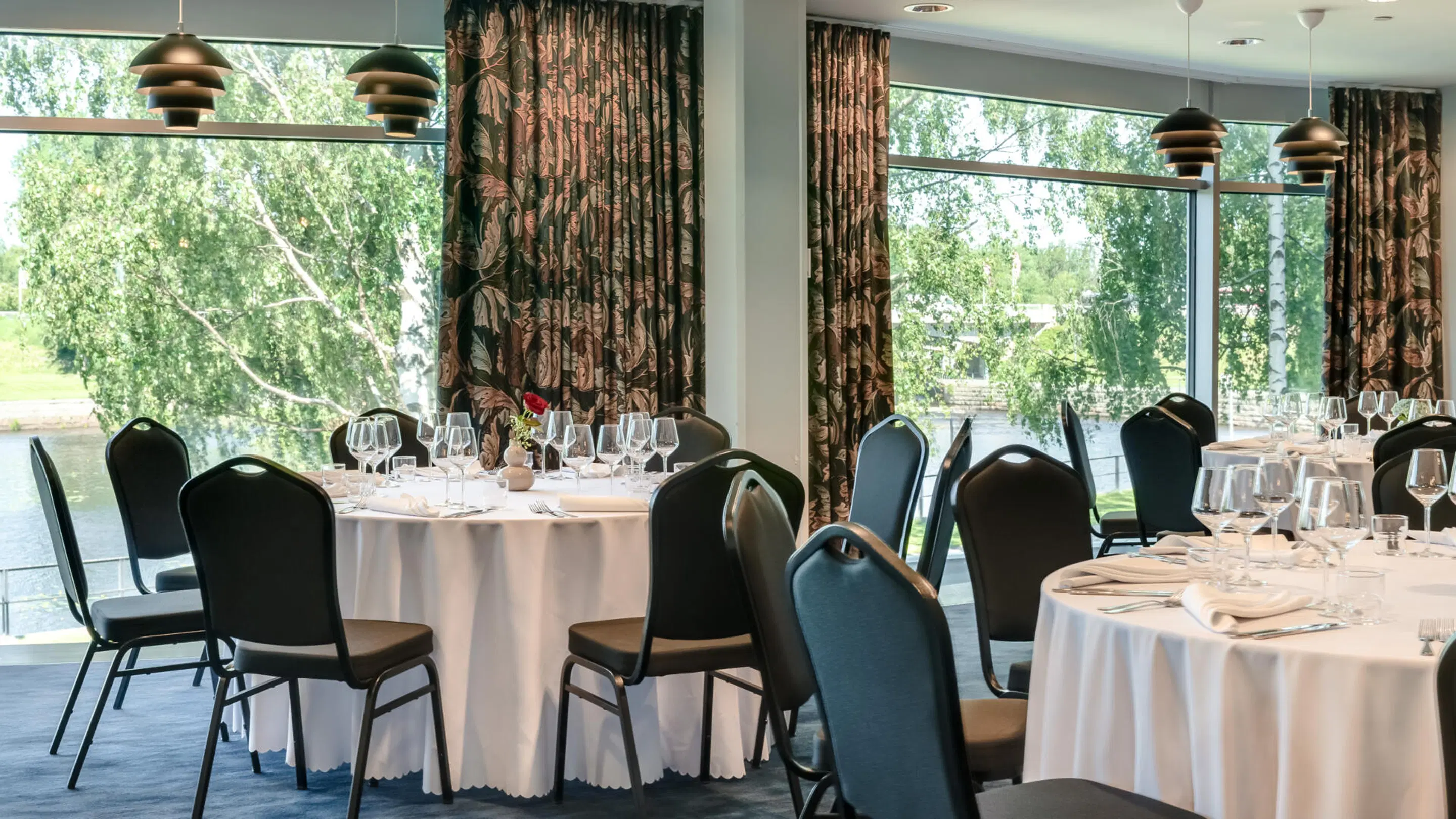 Restaurant dining room with round tables and tall windows overlooking trees. Smooth floor surface and easily movable chairs.