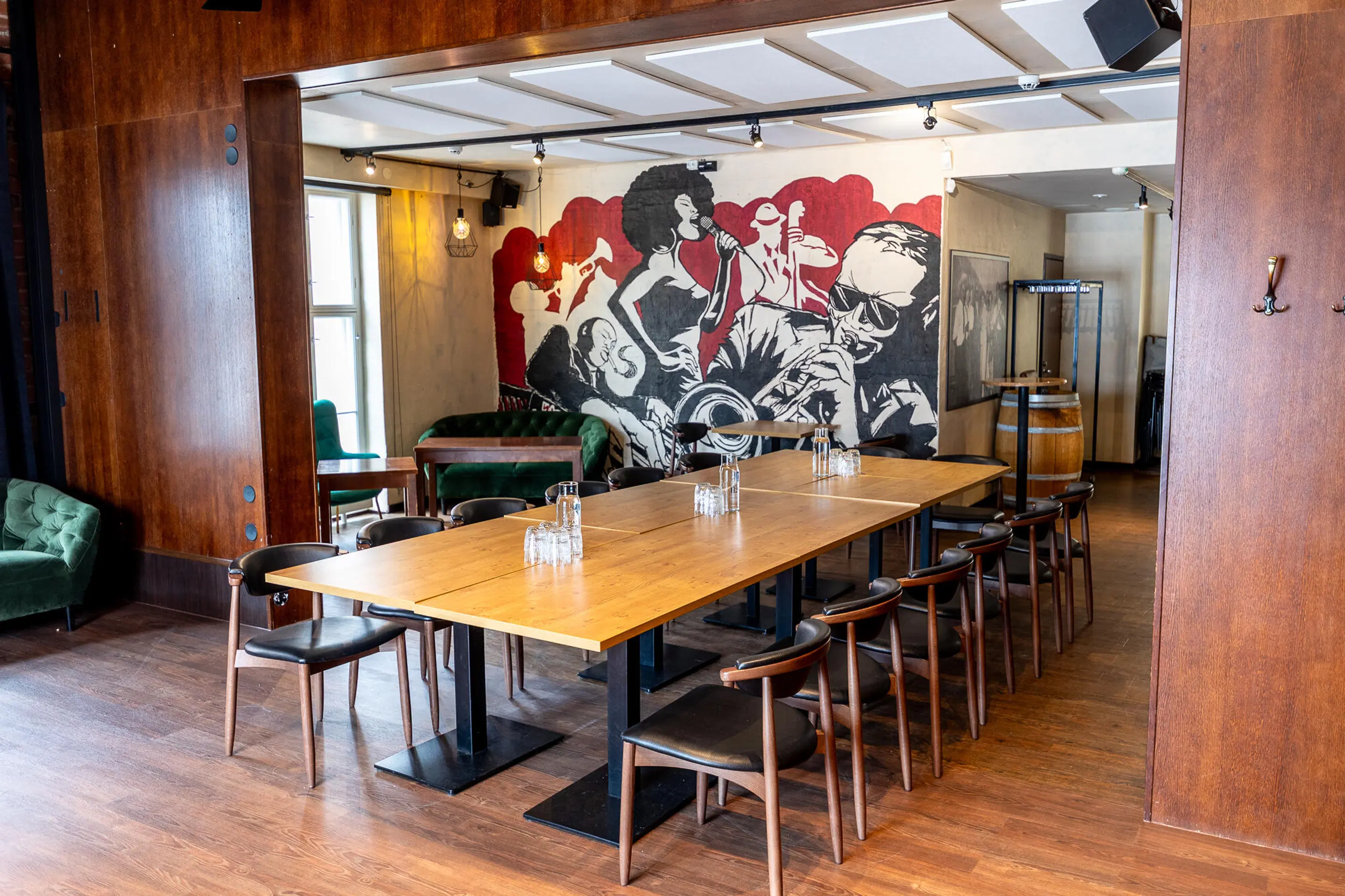 Restaurant interior with wooden table and chairs, green sofa, and music-themed black and white mural with red background. Wooden flooring and good lighting.