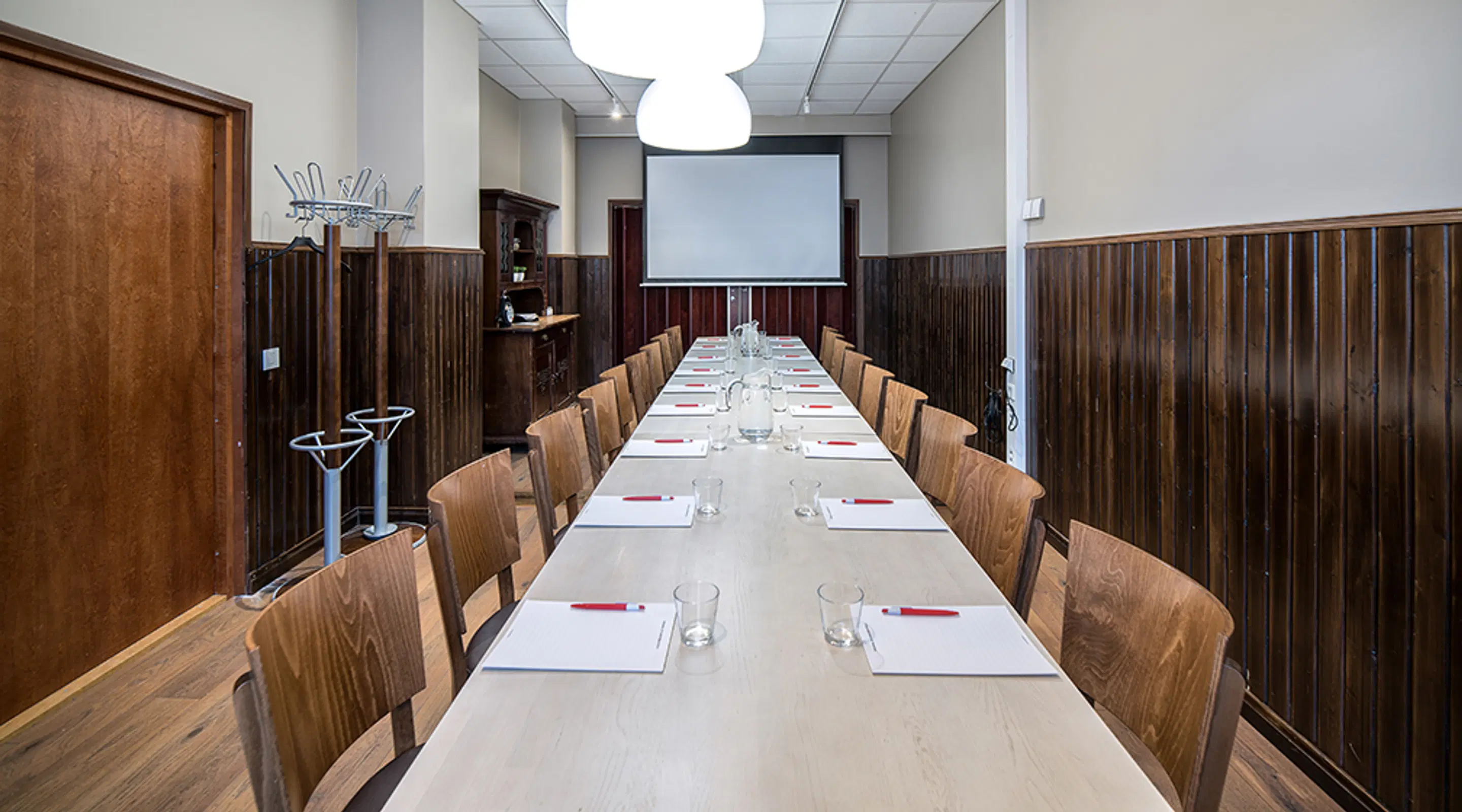 Long meeting room with wooden paneling. White table, wooden chairs, projector screen, glasses and notepads arranged on the table. High ceiling with lighting.