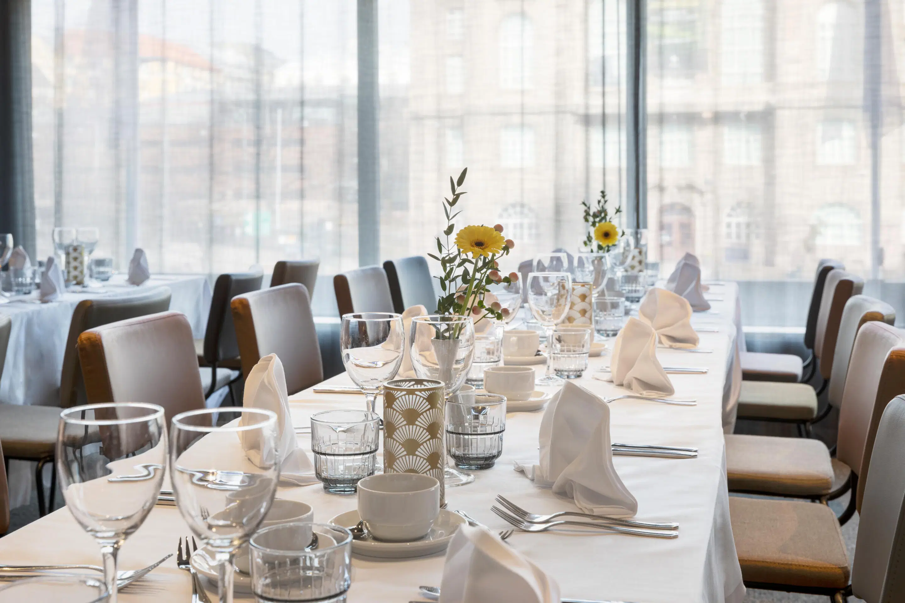Ljus restaurangmatsal med vita dukar, elegant dukning och gula blommor i små vaser. Stora fönster med utsikt.