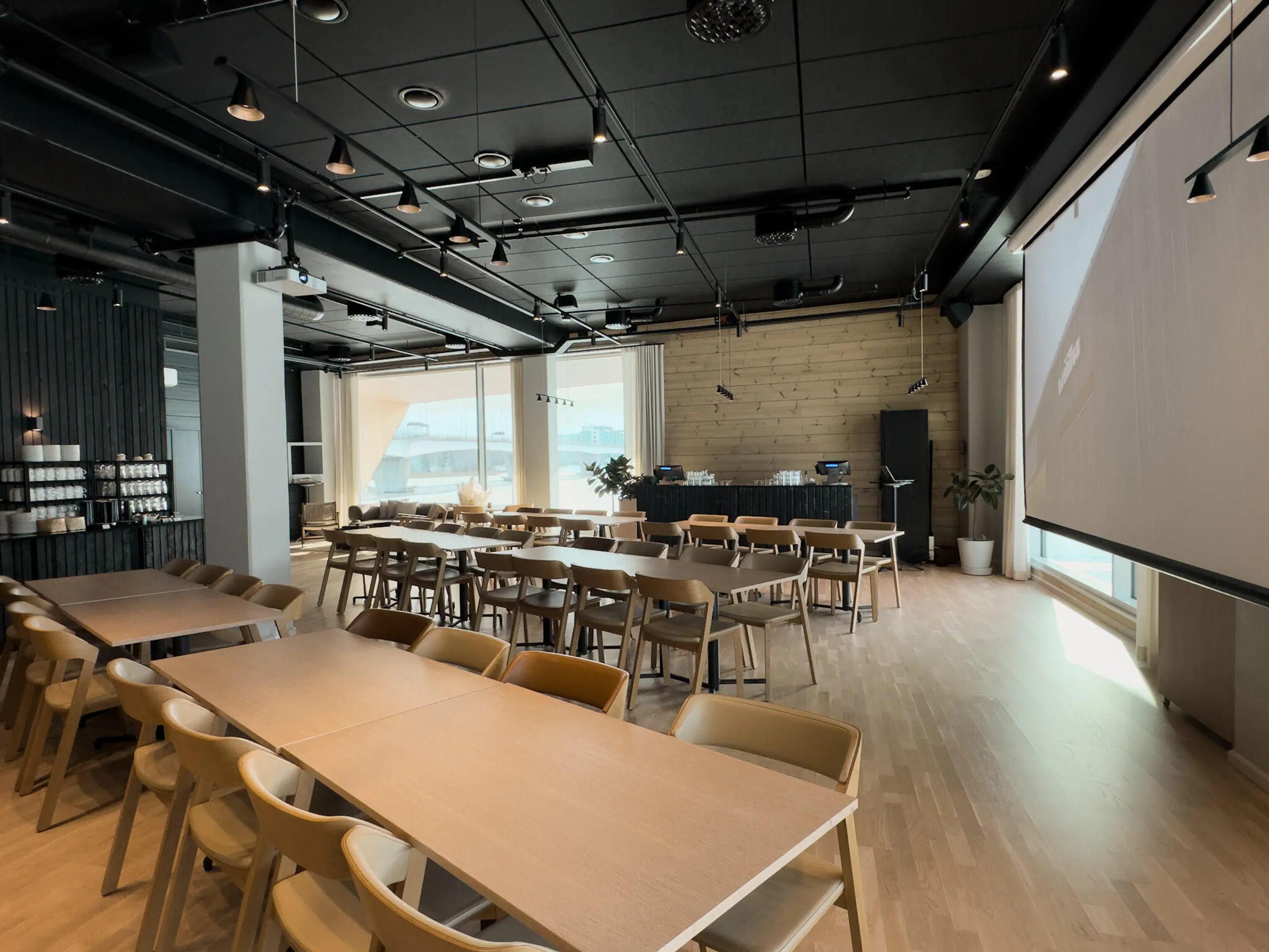 Modern meeting room with wooden tables and chairs. Space features a projector, wooden floor, large windows, and a serving table at the back.