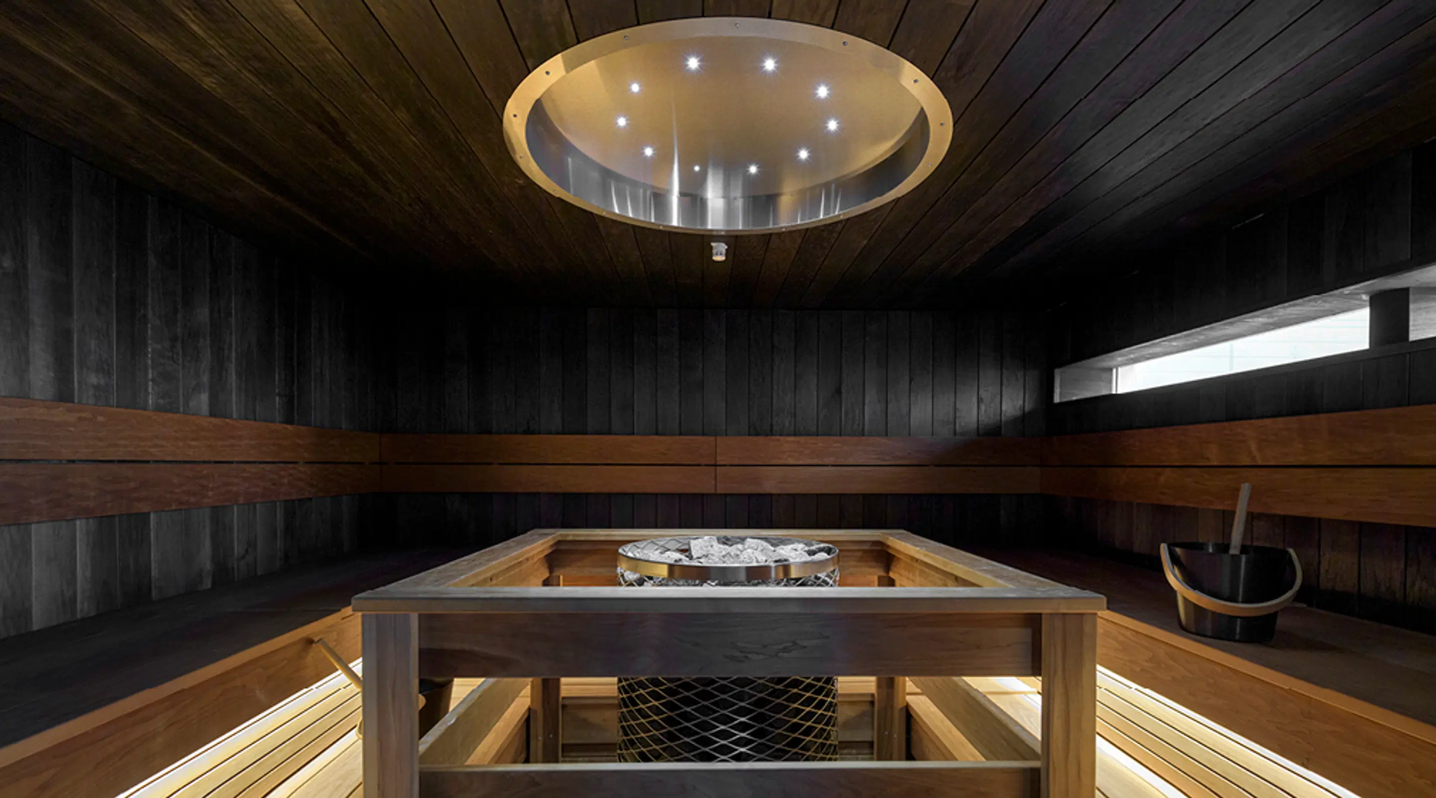 Modern dark sauna with wooden paneling, circular ceiling light, wooden benches, and a heater in the center.