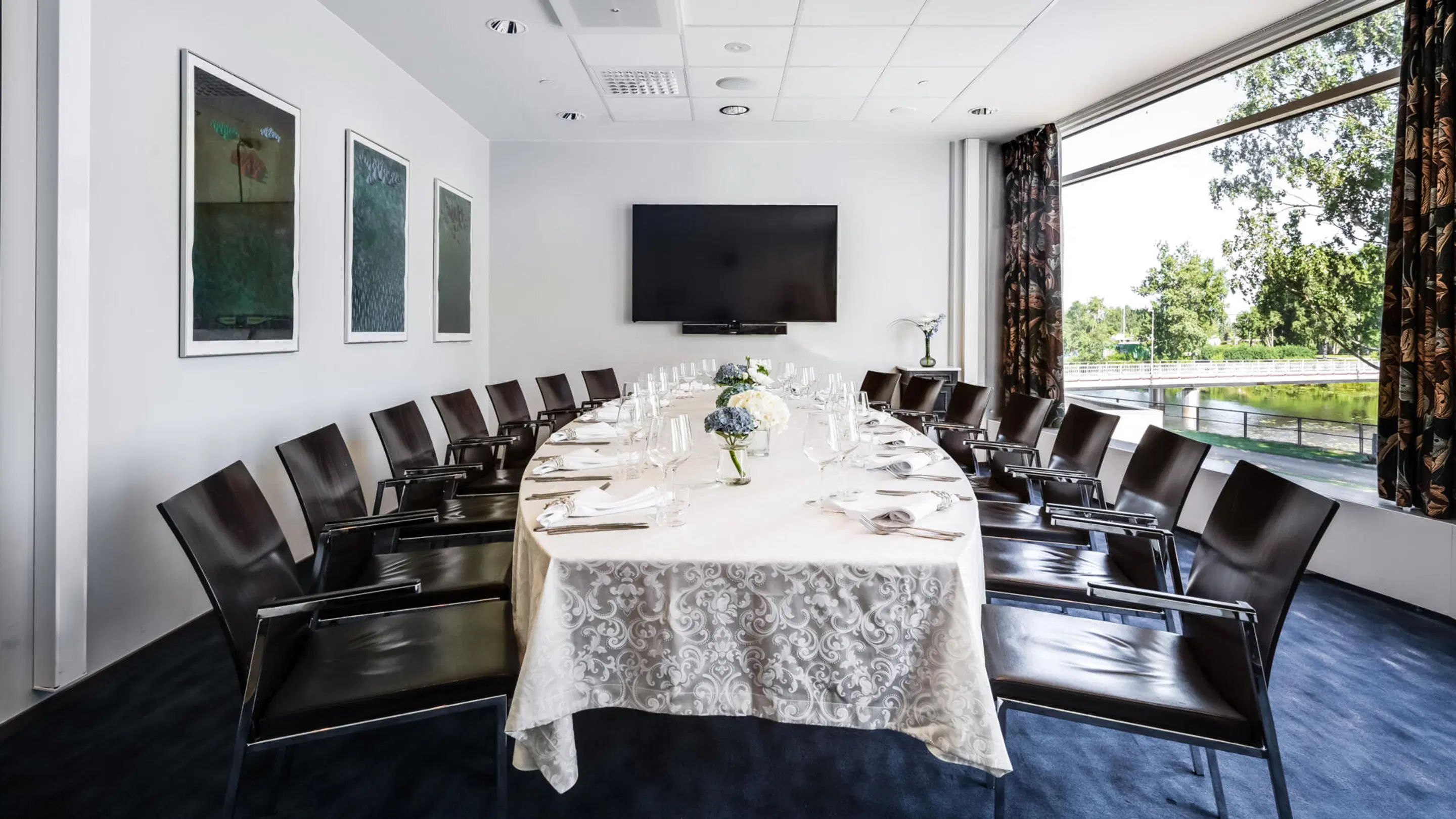 Conference room with a long table covered by white tablecloth, dark chairs, wall-mounted TV, and large windows overlooking a park view with water.