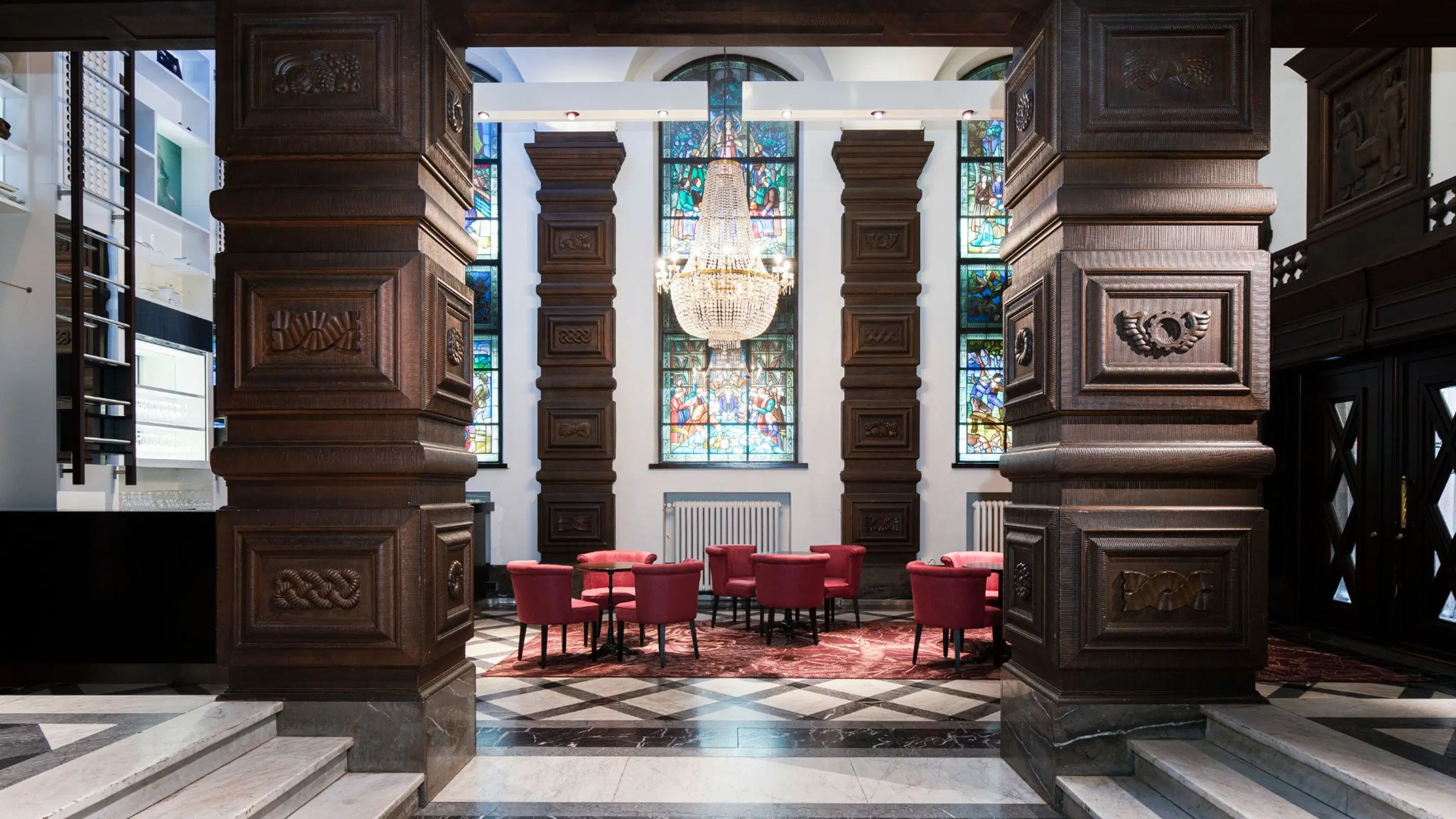 Elegant room with wooden pillars and stained glass windows. Red armchairs on marble floor, crystal chandelier hanging from ceiling. Few steps at entrance.