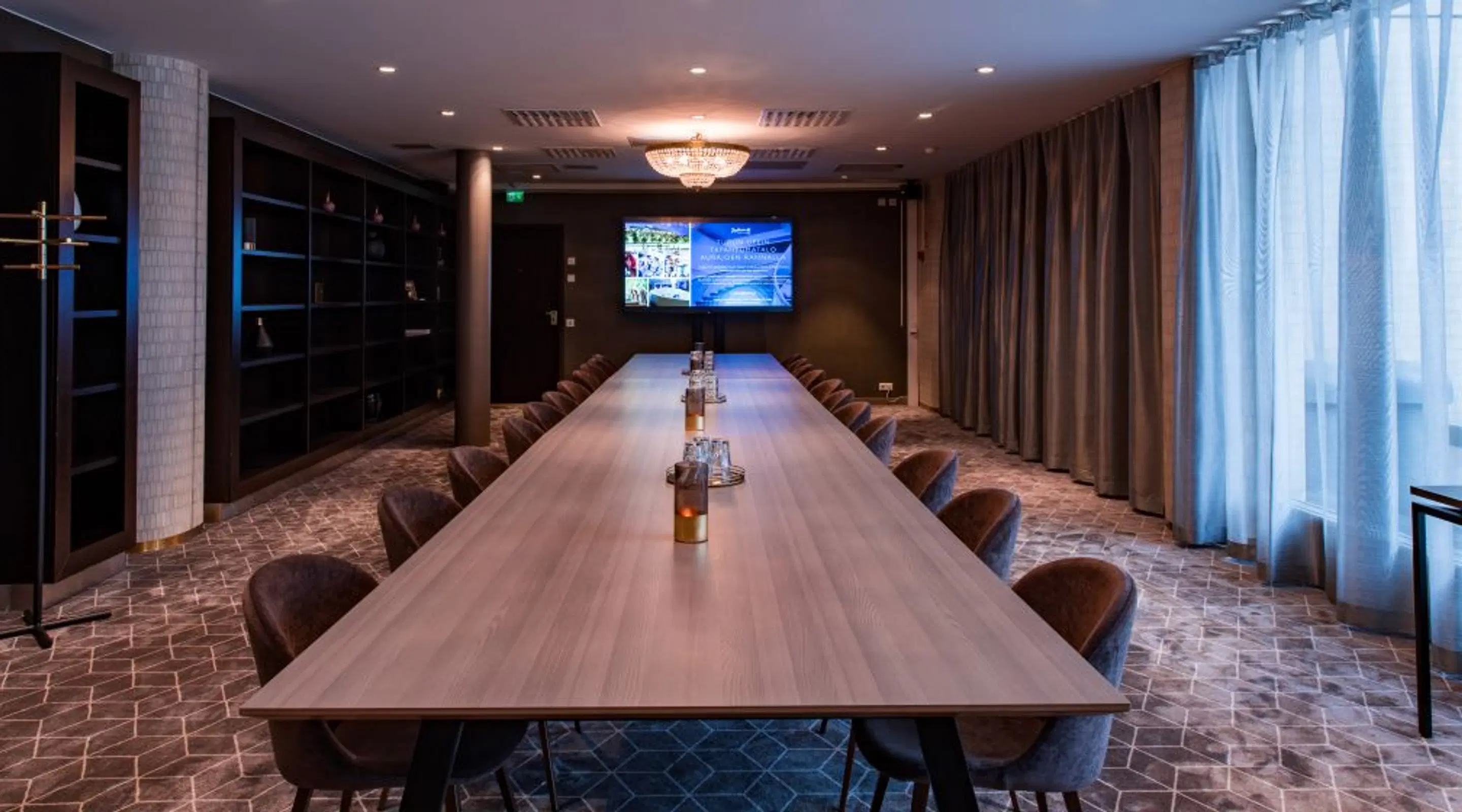 Modern conference room with long wooden table, brown chairs, shelving unit and display screen. Chandelier lighting and natural light from window with curtains.