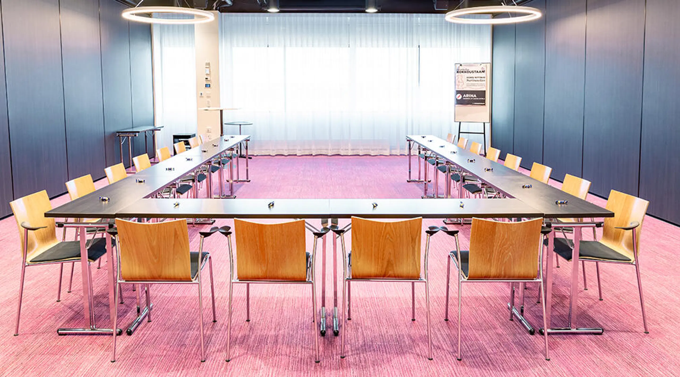 Conference room with U-shaped table arrangement, wooden chairs, pink wall-to-wall carpet and modern ring-shaped lights hanging from the ceiling.