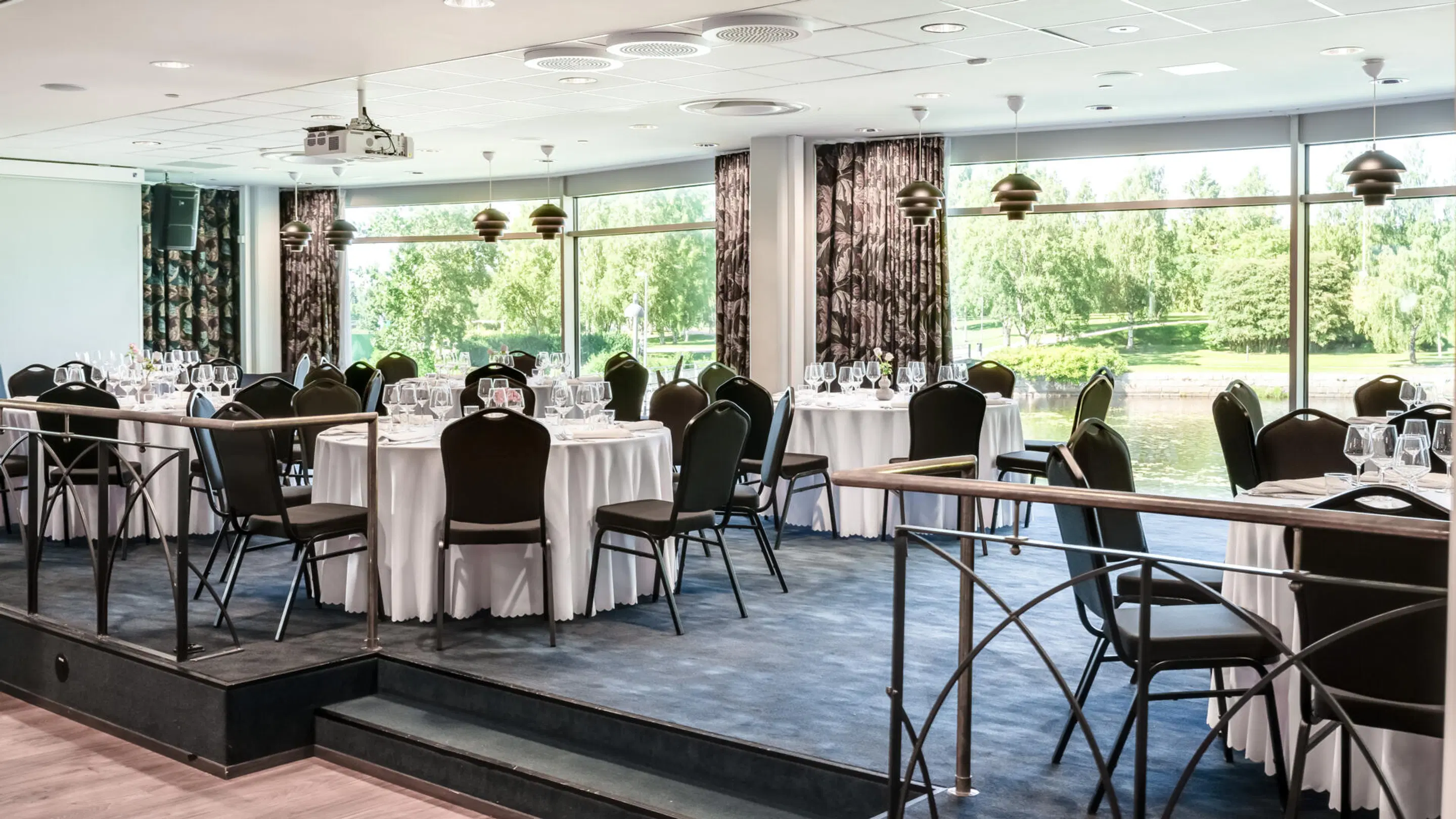 Restaurant dining room with white tables and black chairs. The space features a raised floor and large windows overlooking a park-like landscape with water.