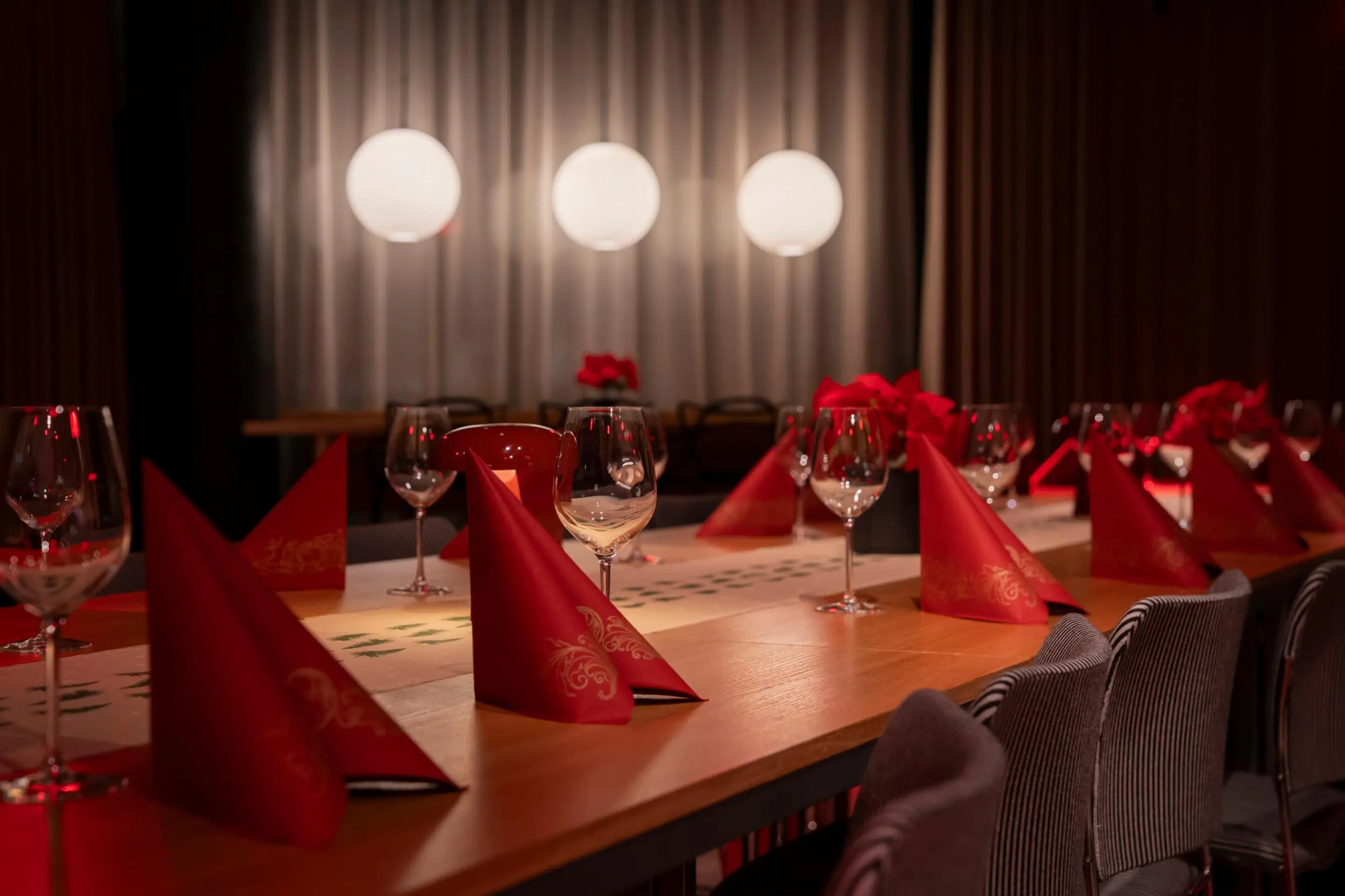 Restaurant table set with red napkins and wine glasses. White curtain and three spherical lights in the background.