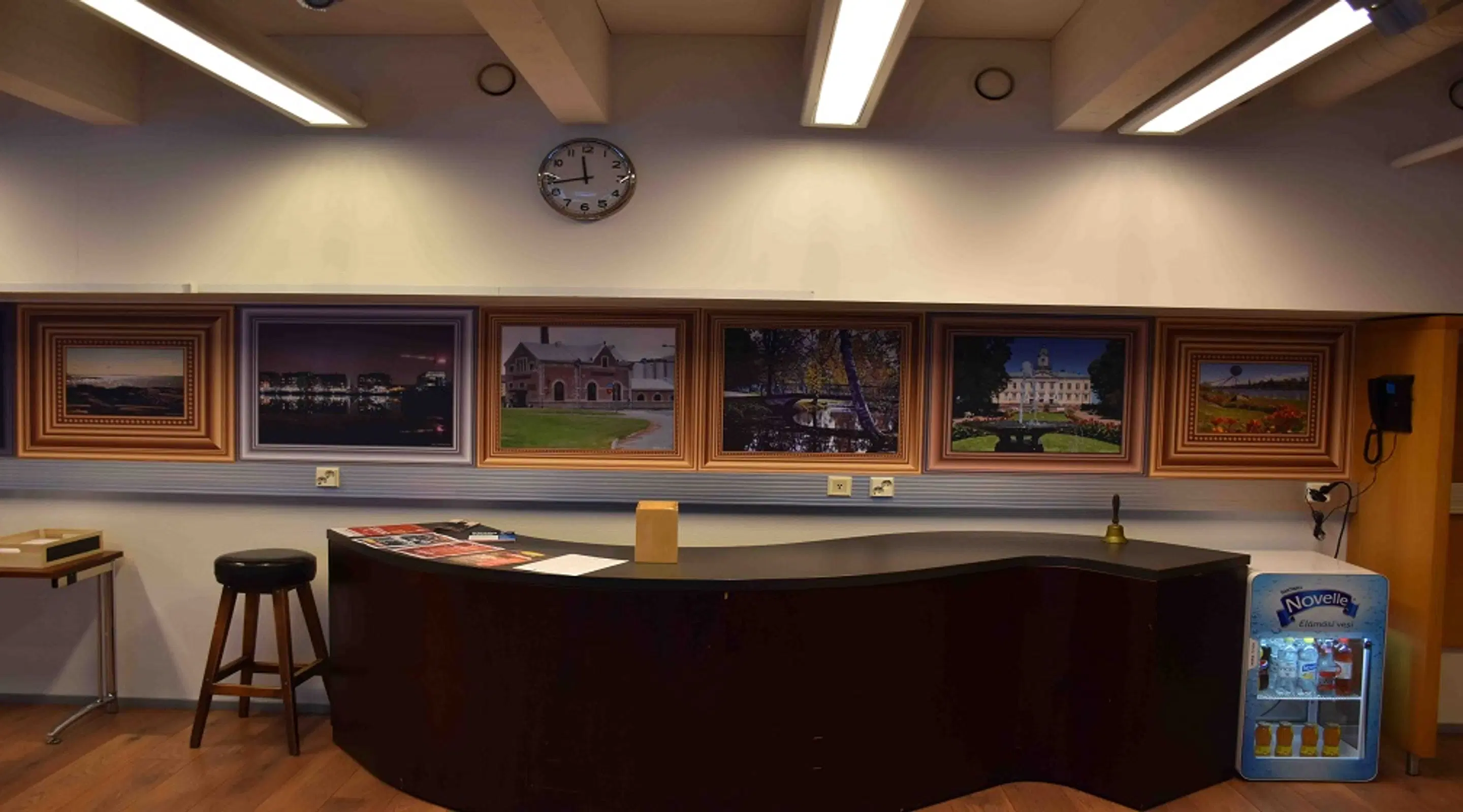Reception desk on wooden floor, wall displays six framed landscape photographs, clock above, tall stool beside and beverage cooler nearby.