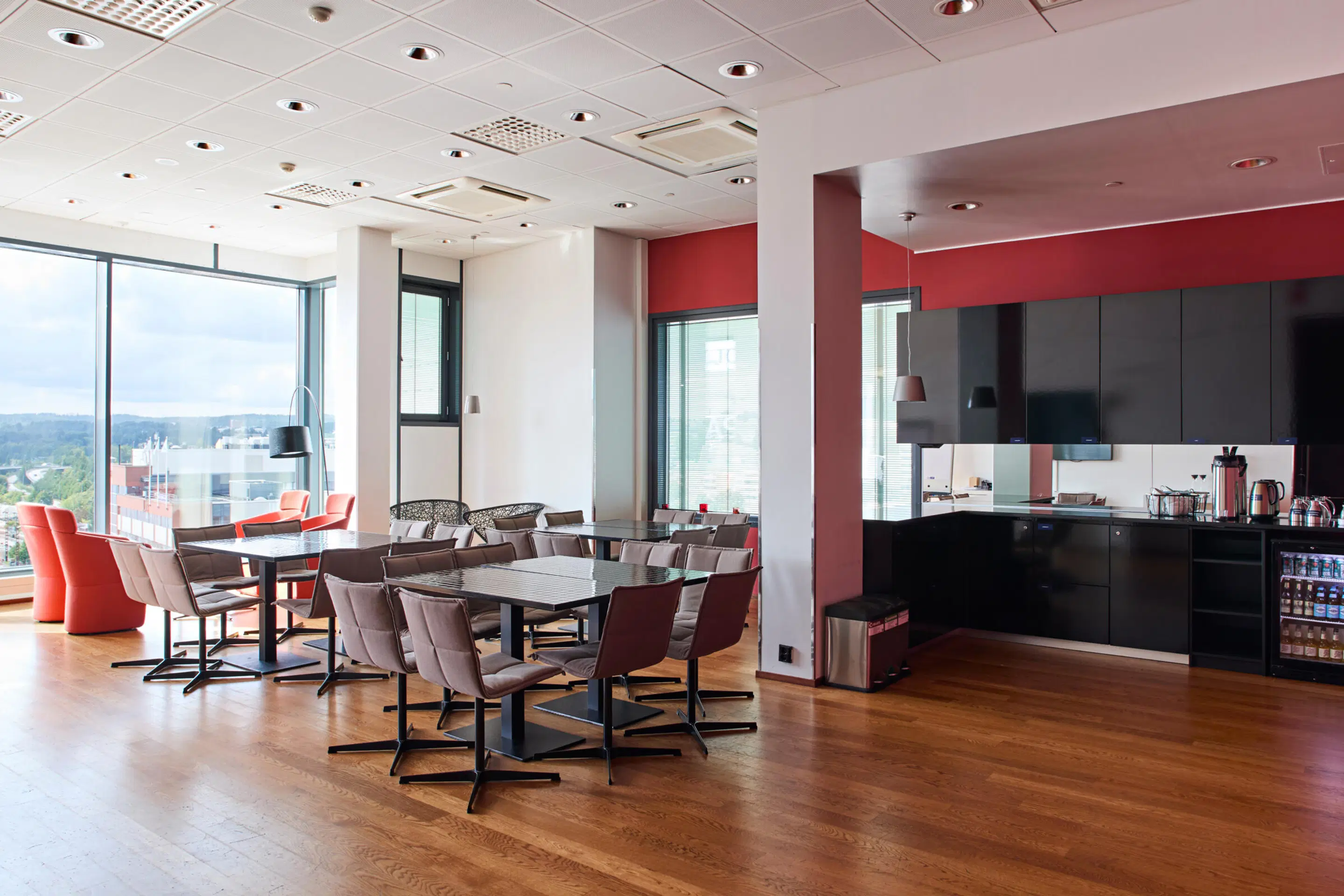 Modern break room with tables and chairs, open kitchen with black cabinets, red wall accents, and large windows overlooking city views.
