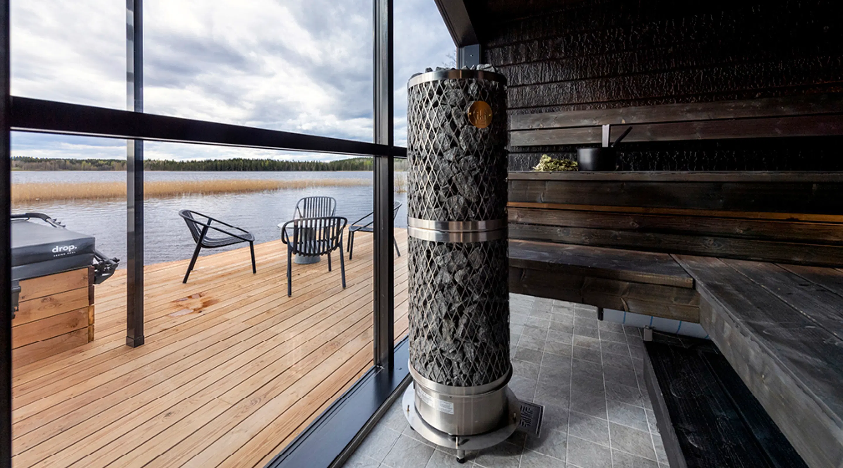 Sauna sisemus ahjuga, klaasseina taga terrass järvevaatega ja toolidega. Hall pilvine taevas üleval.