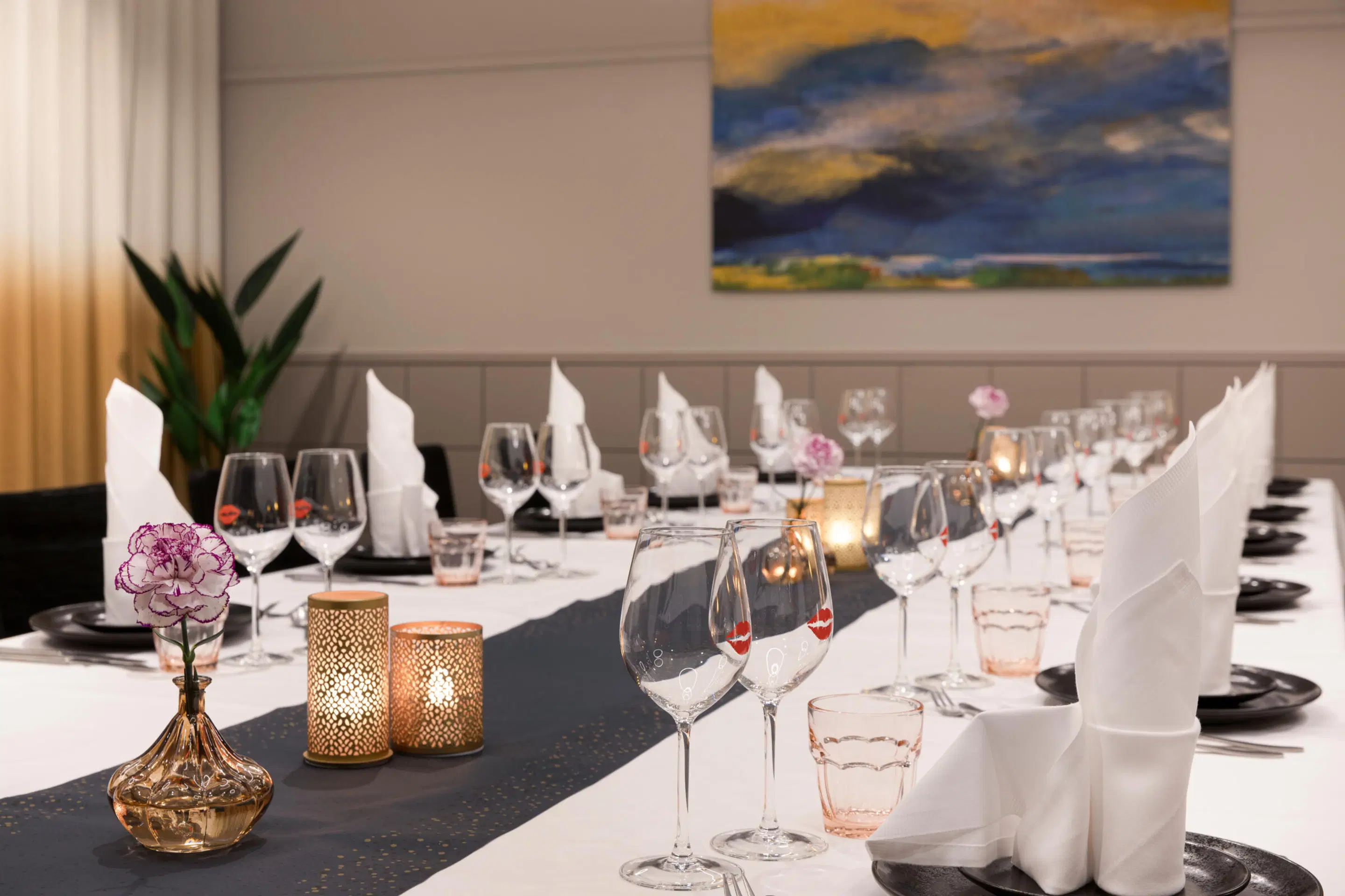 Elegant dukat långbord i en restaurang. Vit duk med svarta tallrikar och vinglas. Bordet har ljus och blomsterdekorationer. En blåtonad konstmålning syns i bakgrunden.