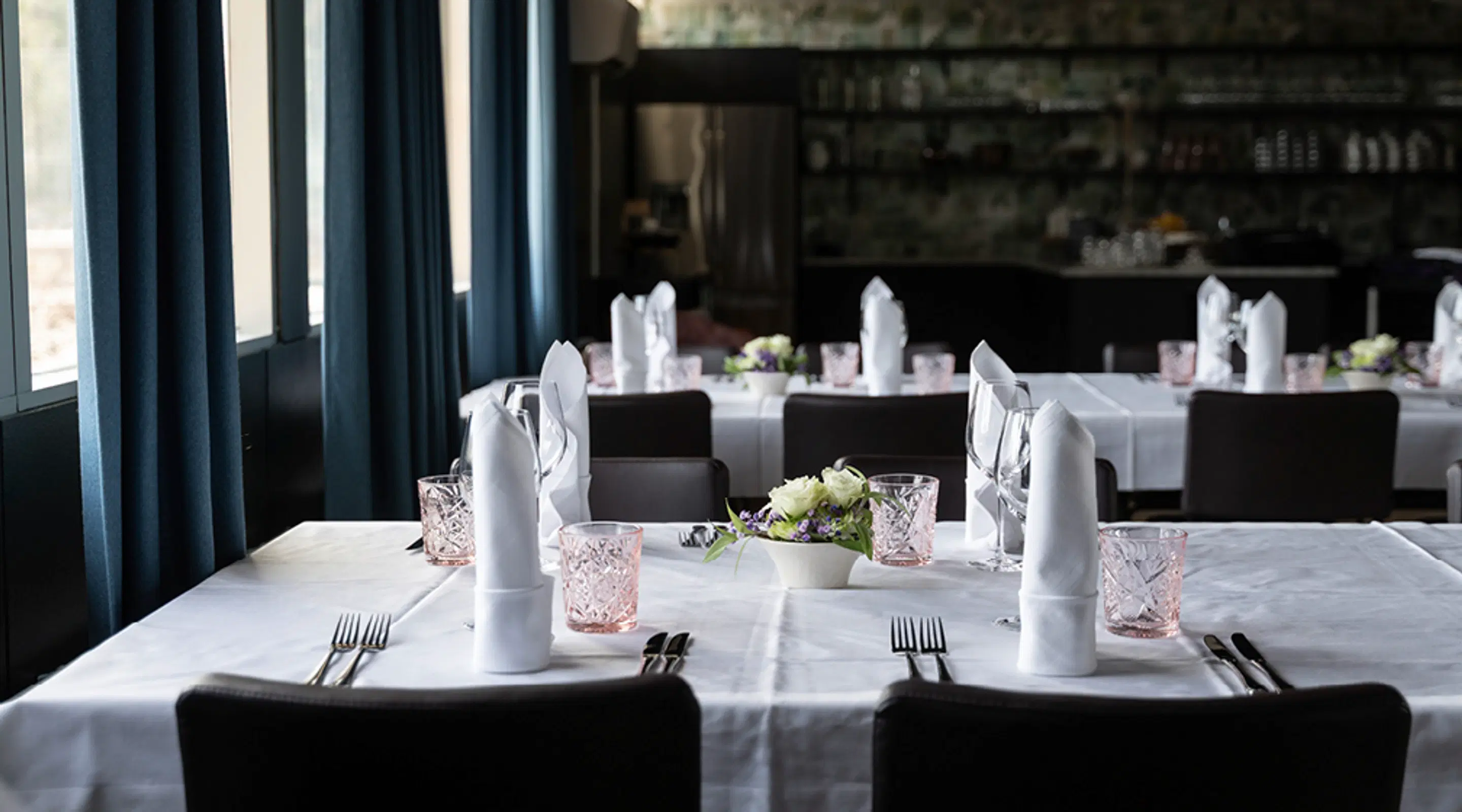 Elegantly set restaurant tables with white tablecloths, folded napkins, pink drinking glasses and dark chairs in front of blue curtains.