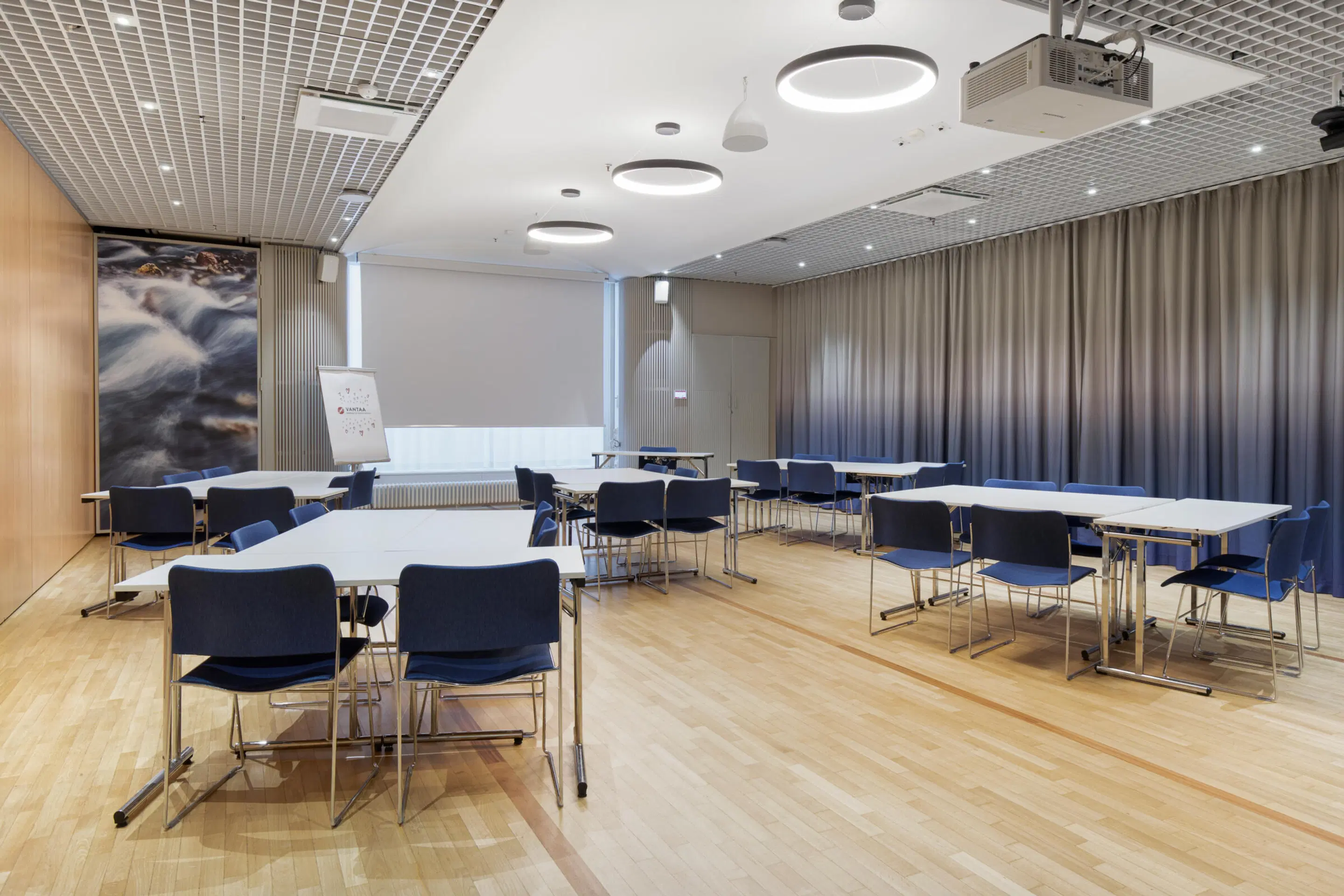 Modern meeting room with white tables and blue chairs arranged on wooden floor. Round ceiling lights, gray curtains, and water rapids image on wall.
