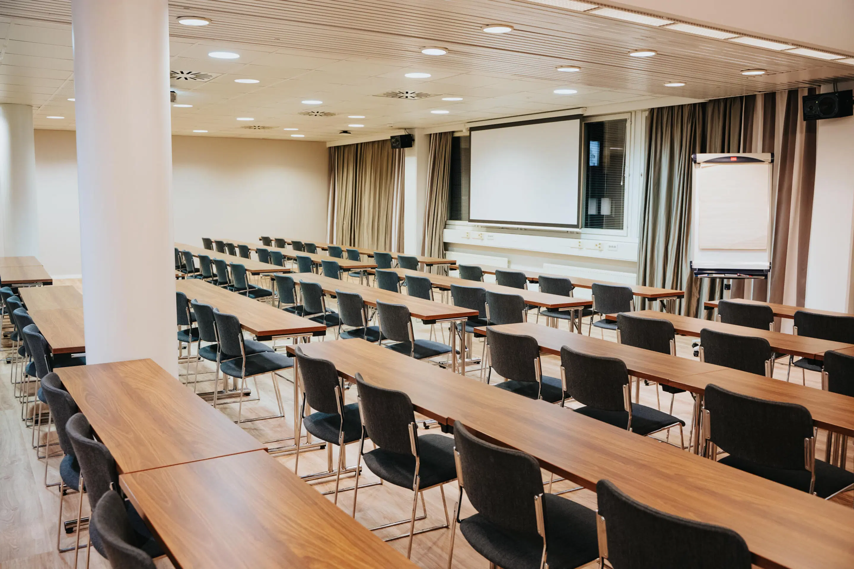 Bright conference room with long wooden tables, black chairs and a presentation screen. The room has a level floor and good lighting.