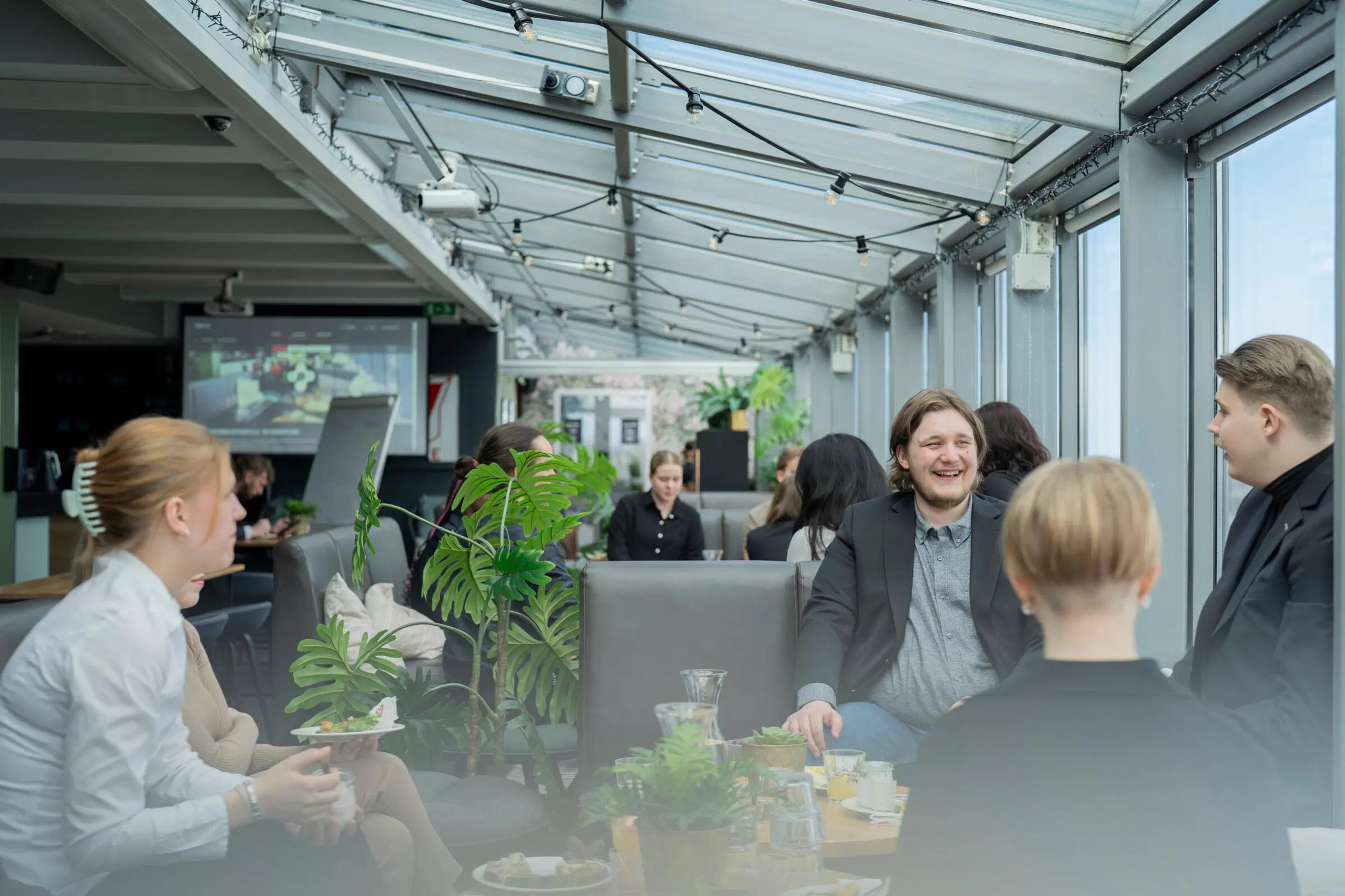 A group of people conversing in a glass-roofed café or restaurant. Tables feature potted plants and drinks. The space is bright with ample natural light.