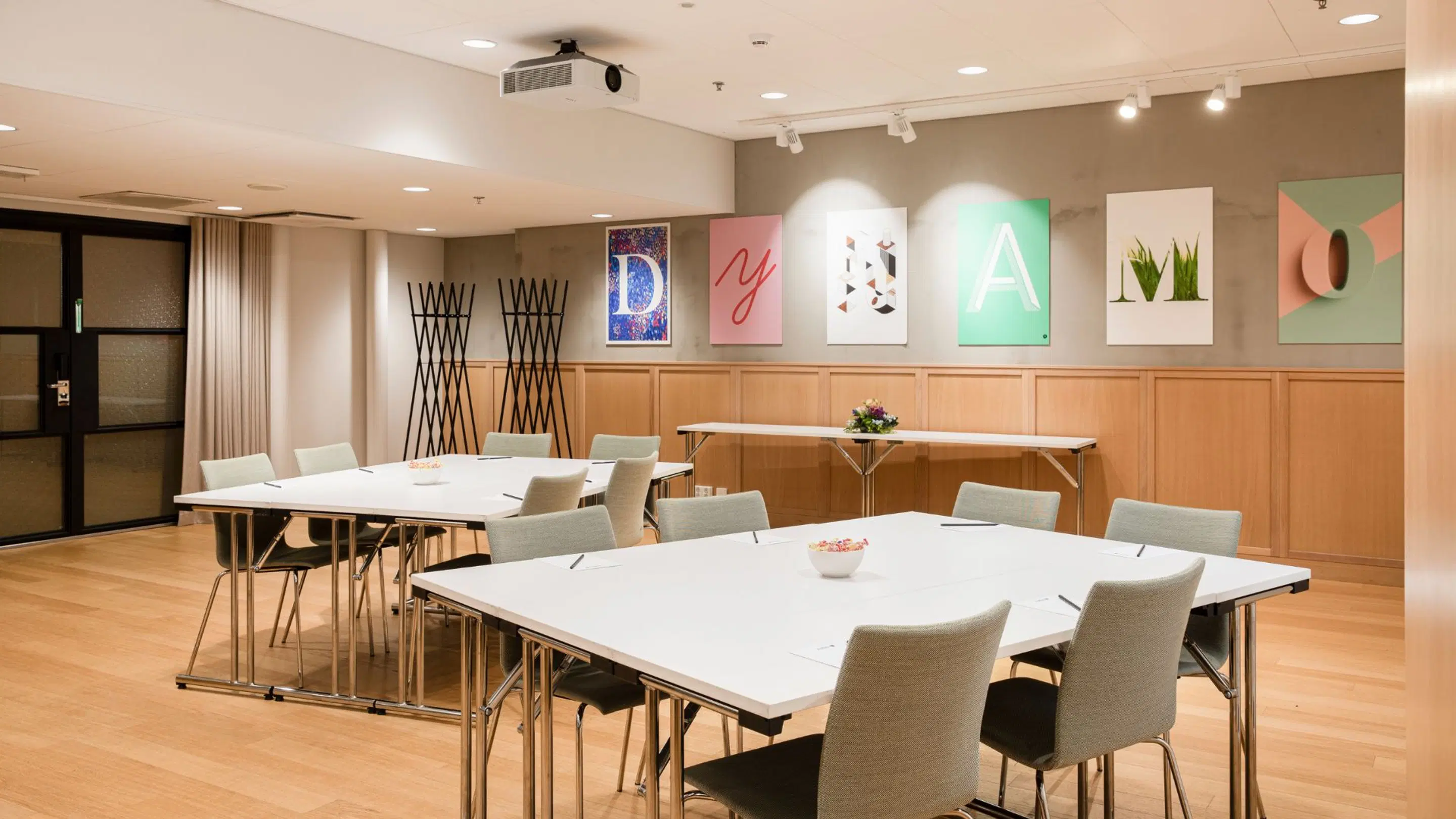 Modern meeting room with two white tables, light gray chairs and art on the wall. Wooden floor and spot lighting.