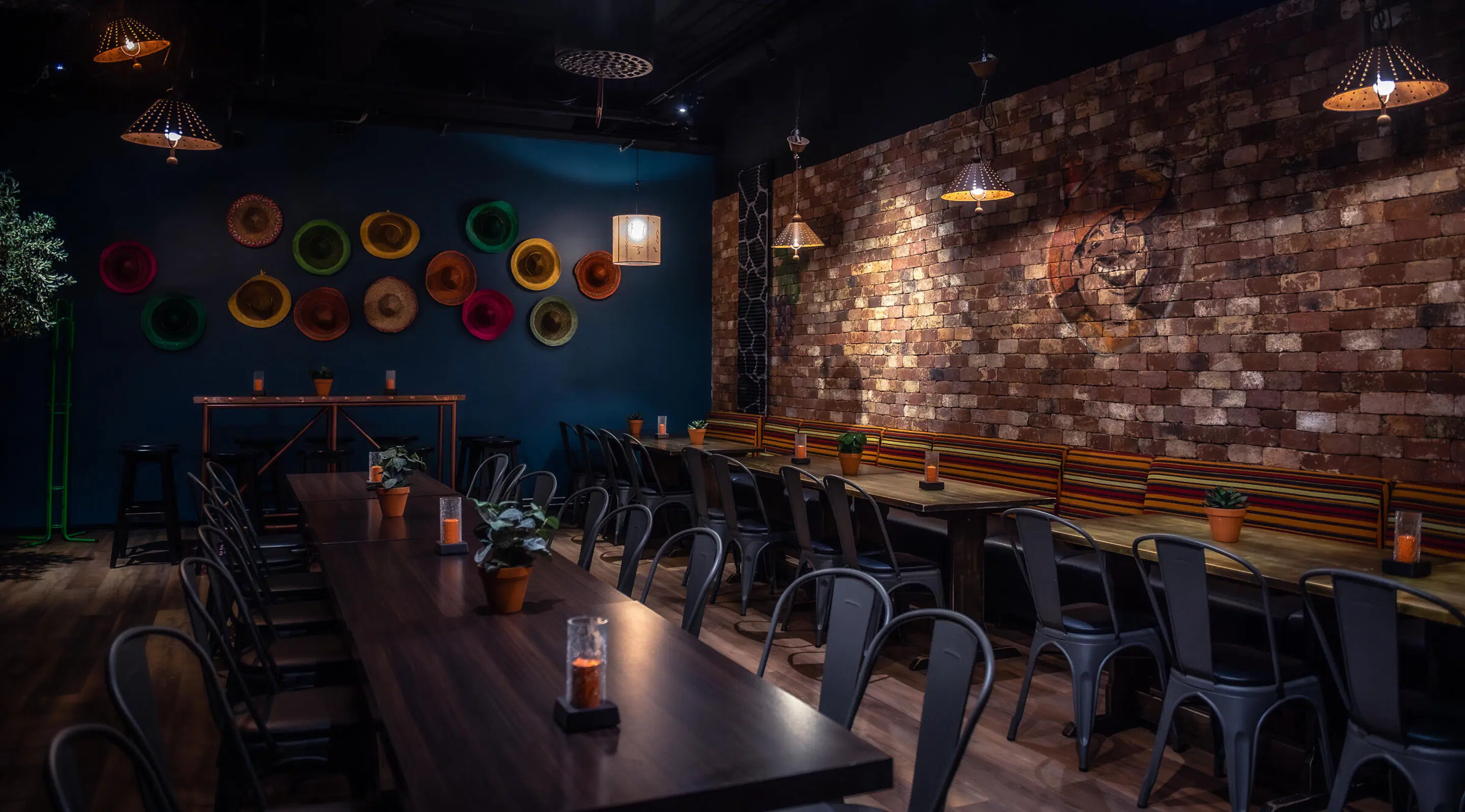 Atmospheric restaurant with dark blue wall decorated with colorful hats, brick wall, and black metal chairs around wooden tables. Ceiling lamps and wooden floor.