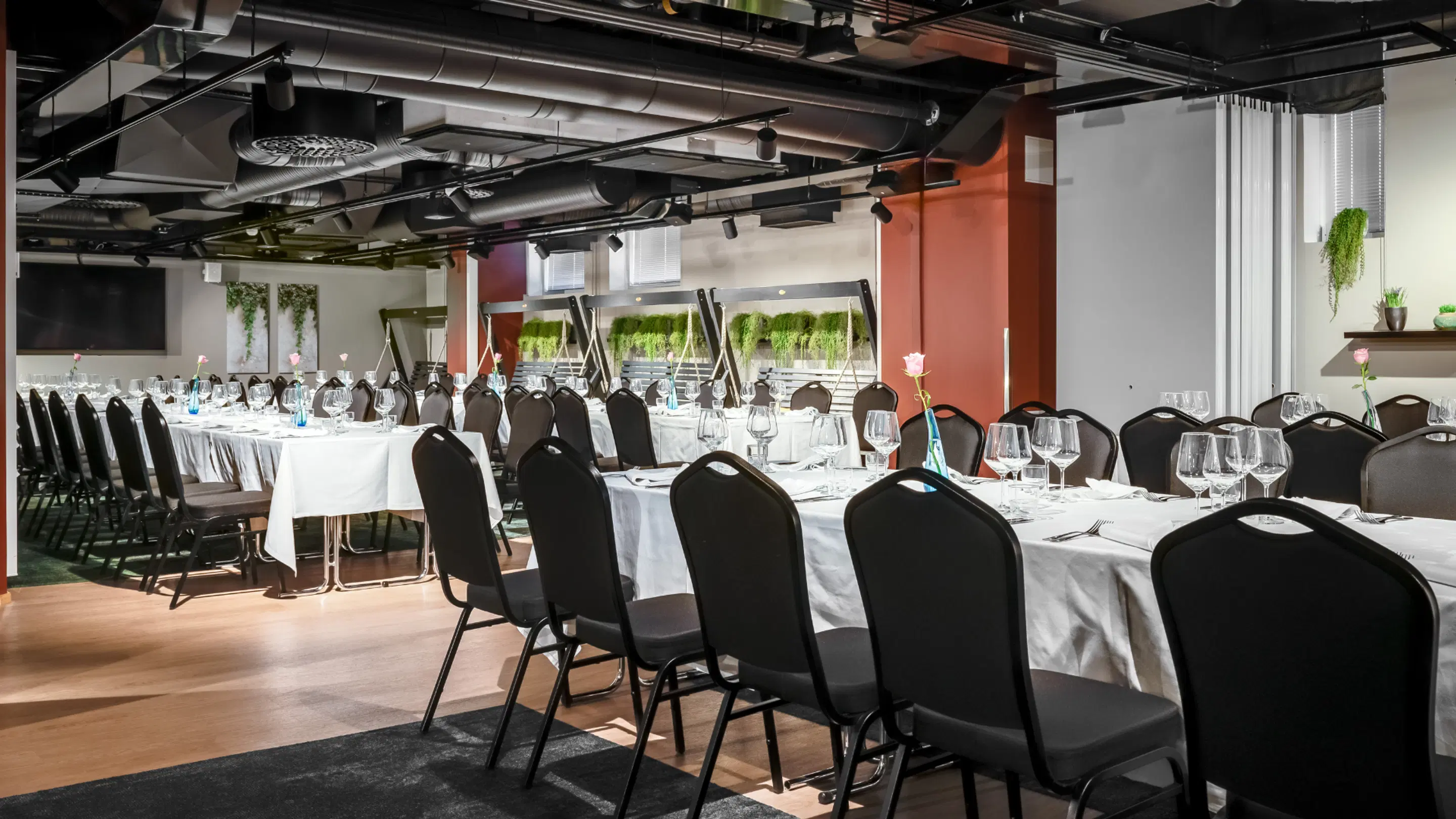 Modern event space with set tables. Long tables with white tablecloths, wine glasses and roses. Room features level wooden floor and wall greenery.