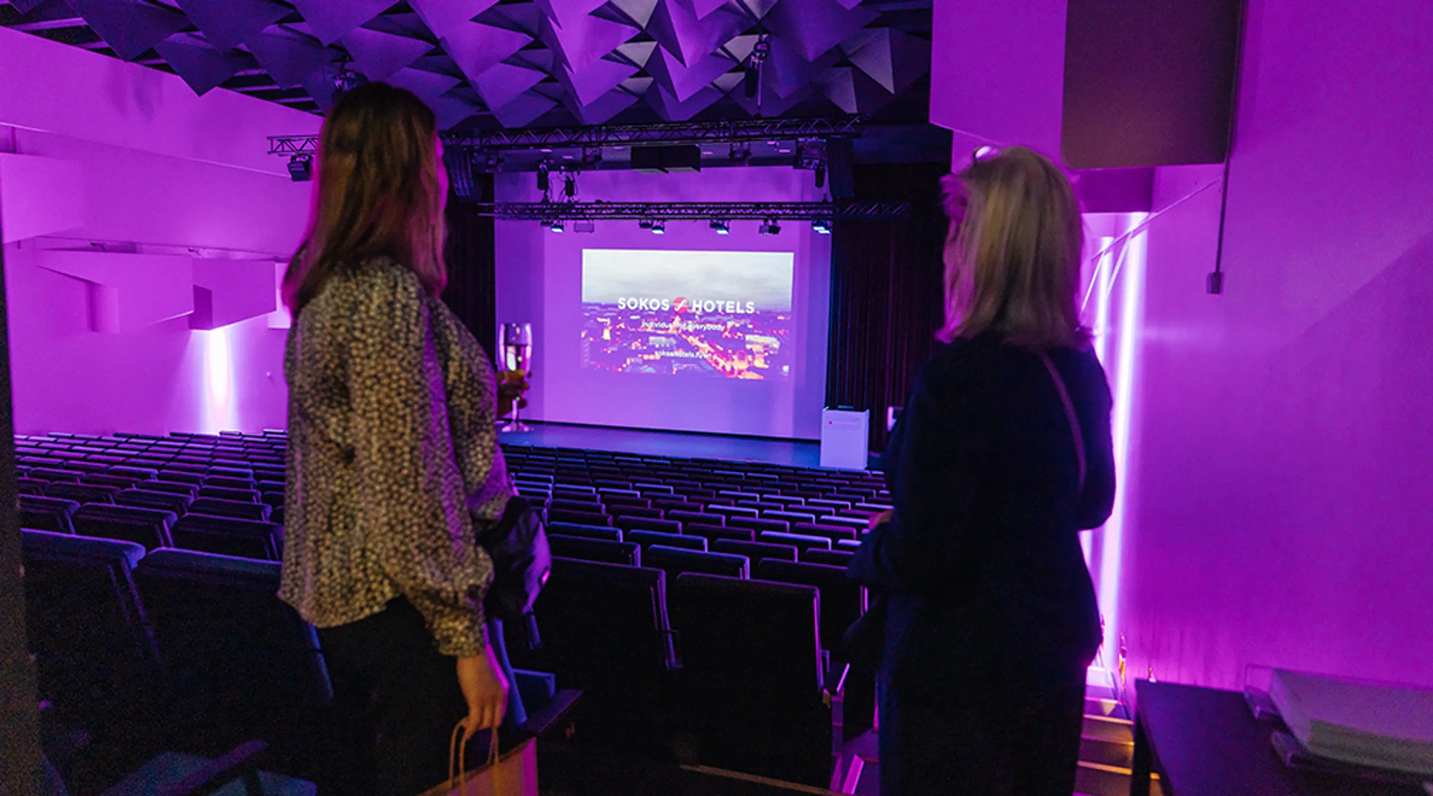Kaksi henkilöä seisoo purppuranvärisesti valaistussa auditoriossa ja katsoo lavaa ja valkokangasta. Penkit ovat porrastetuissa riveissä. Sivulla on portaita.
