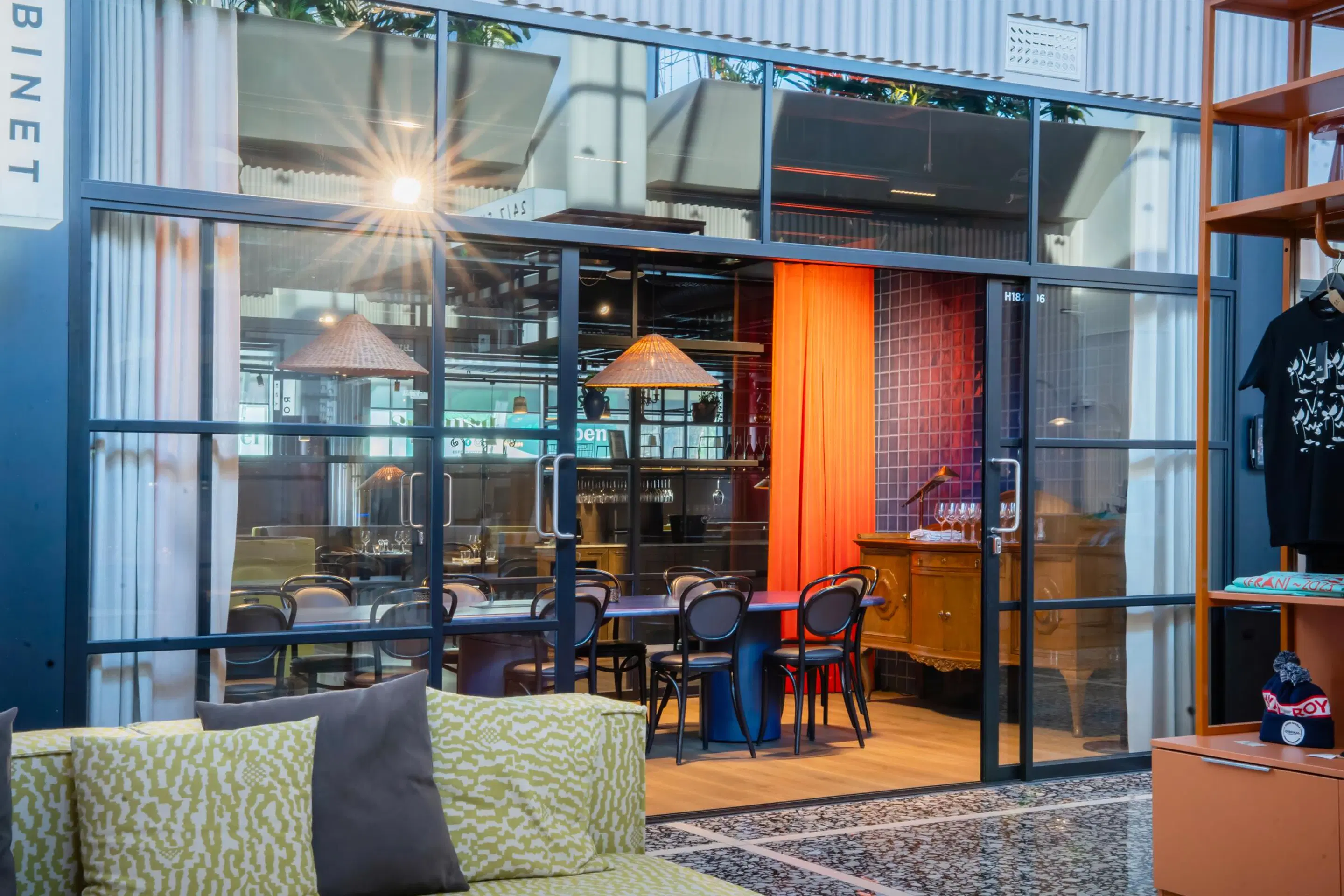 Glass-walled meeting room in restaurant with dark chairs, wooden cabinet and orange curtain. Viewed from lounge area with patterned cushions.
