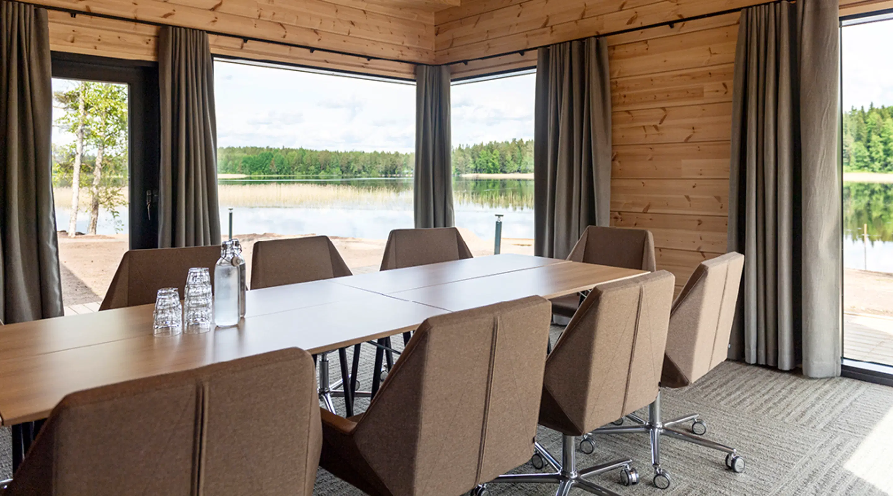 Mötesrum med träpanel och stora fönster med utsikt över sjön och skogslandskapet. Bruna kontorsstolar med hjul runt bordet.