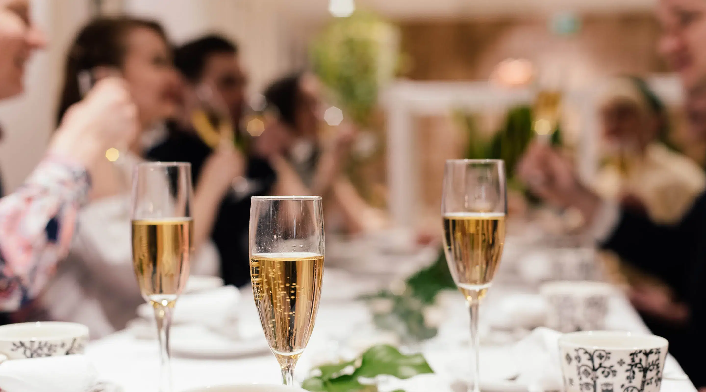Three champagne flutes on white tablecloth, with blurred party guests and green decorations in the background.