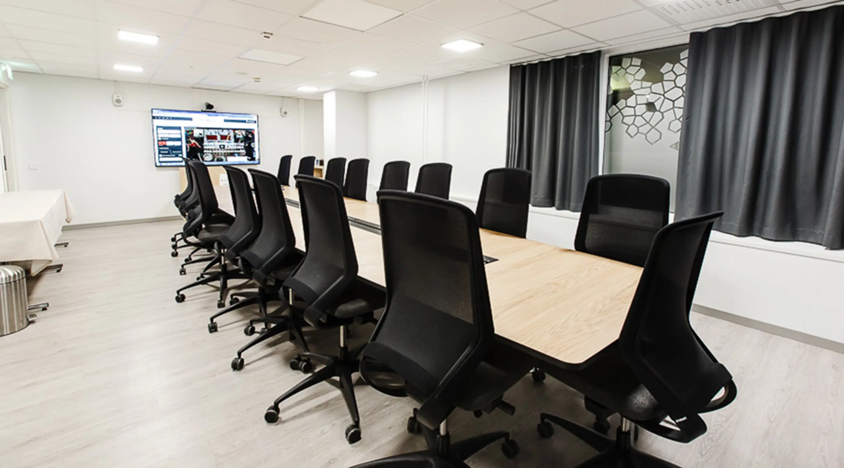 Bright meeting room with a long wooden table and black office chairs. Large screen on the wall and grey curtains. Floor is light laminate.