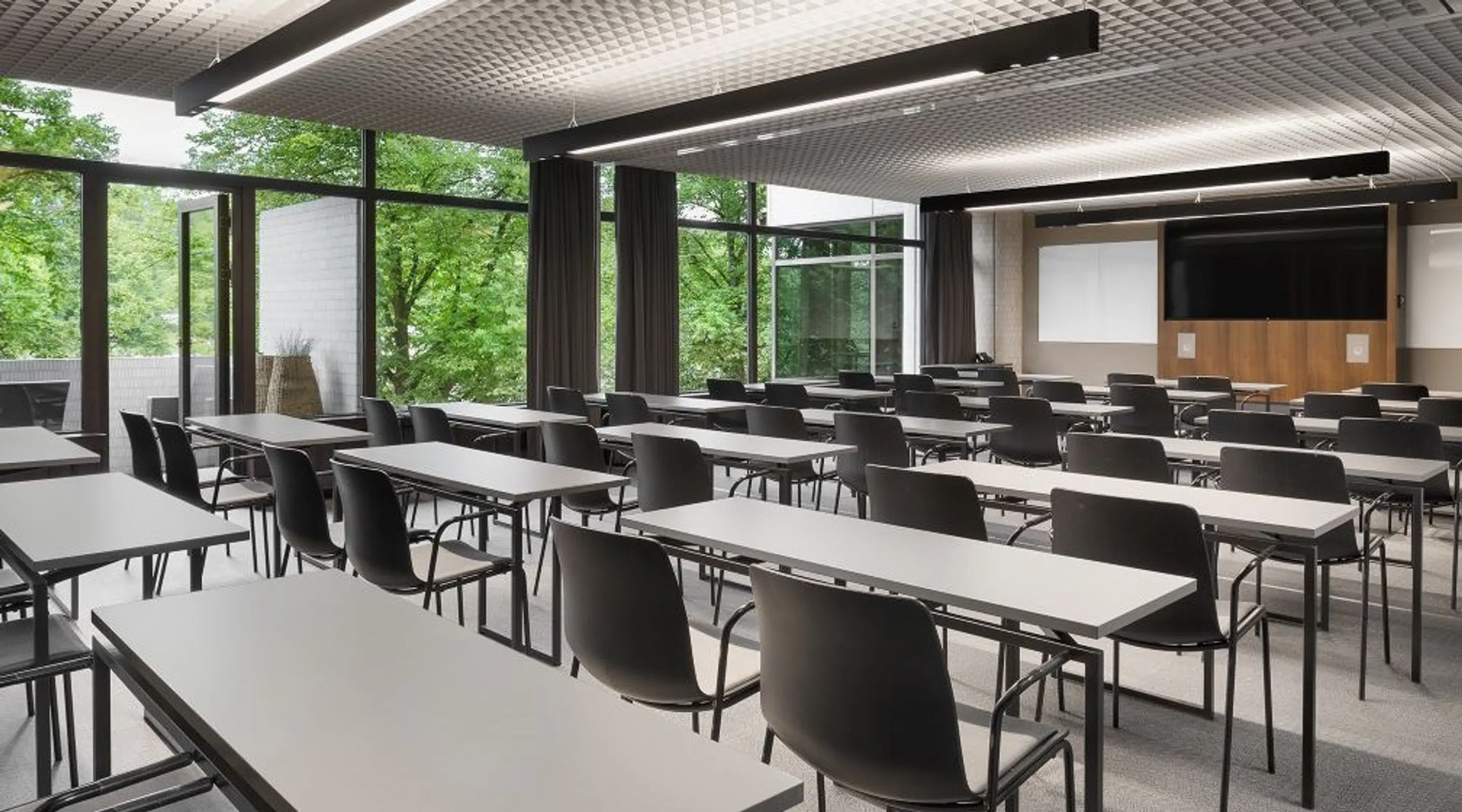 Modern classroom with white tables and black chairs arranged in rows. Large windows overlook green trees. Projector screen and display at front.