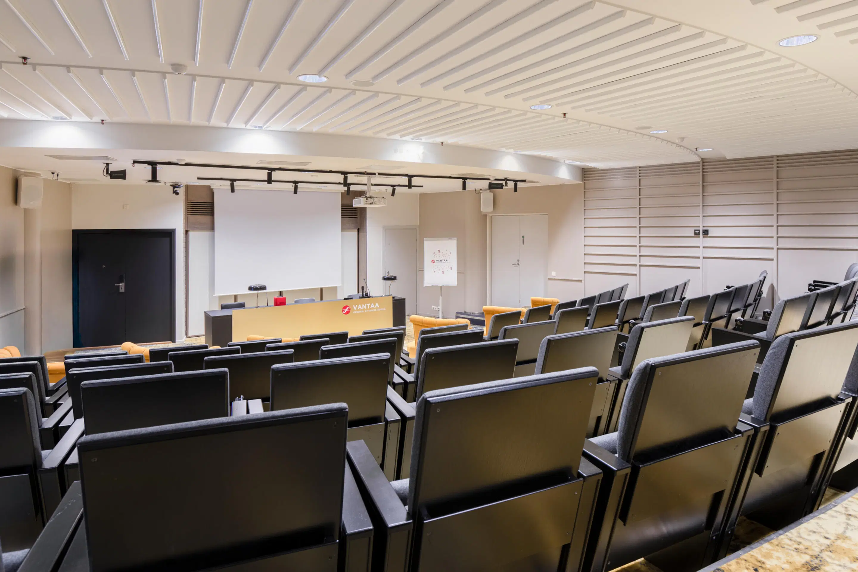 Moderni auditorio järjestetyllä istuintilalla, valkoisella projektorikankaalla ja esiintymispöydällä. Tilavuuden katossa on kiinnitettyjä valaisimia.