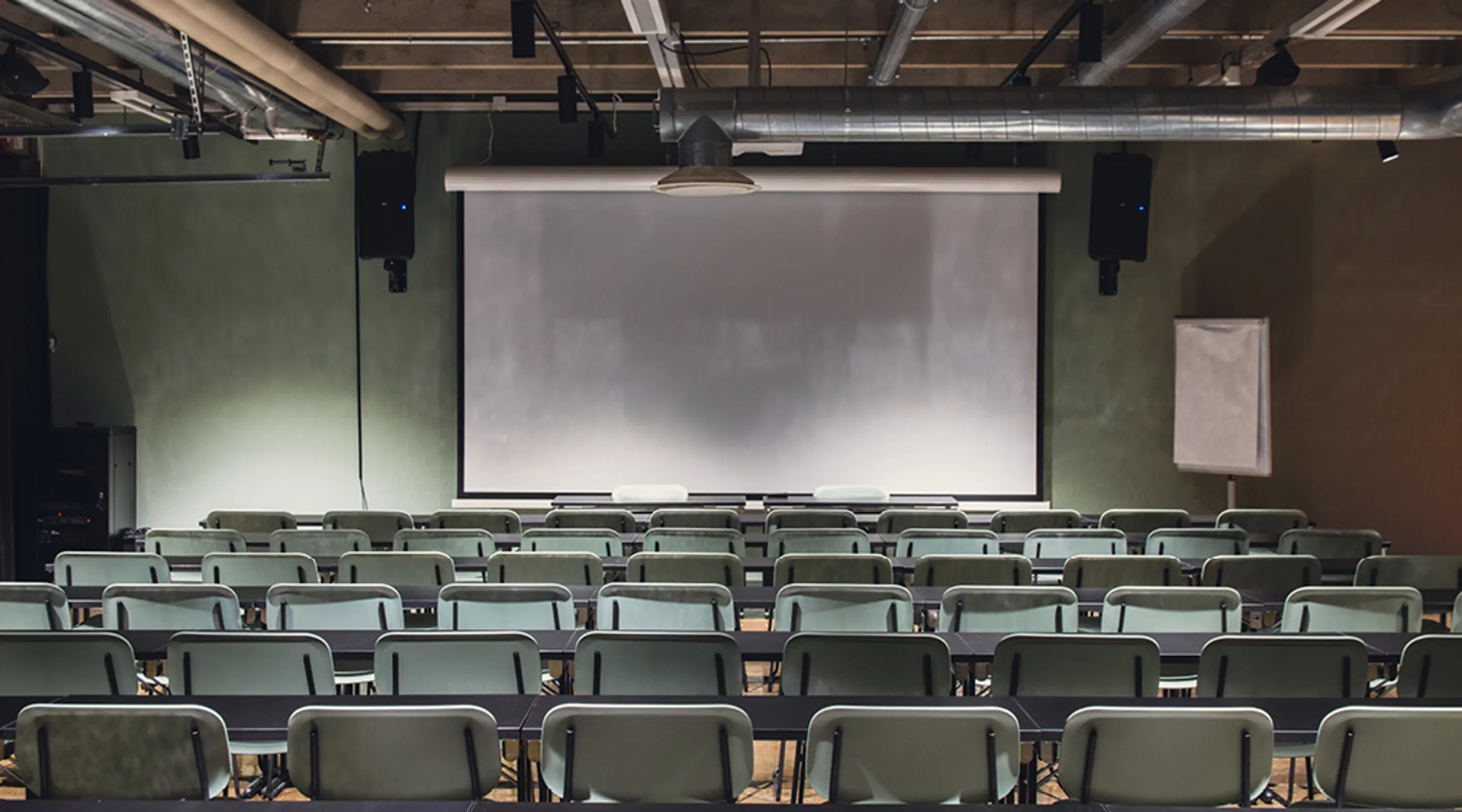 Room arranged in classroom style with empty chairs and a projection screen. Speakers on the wall.