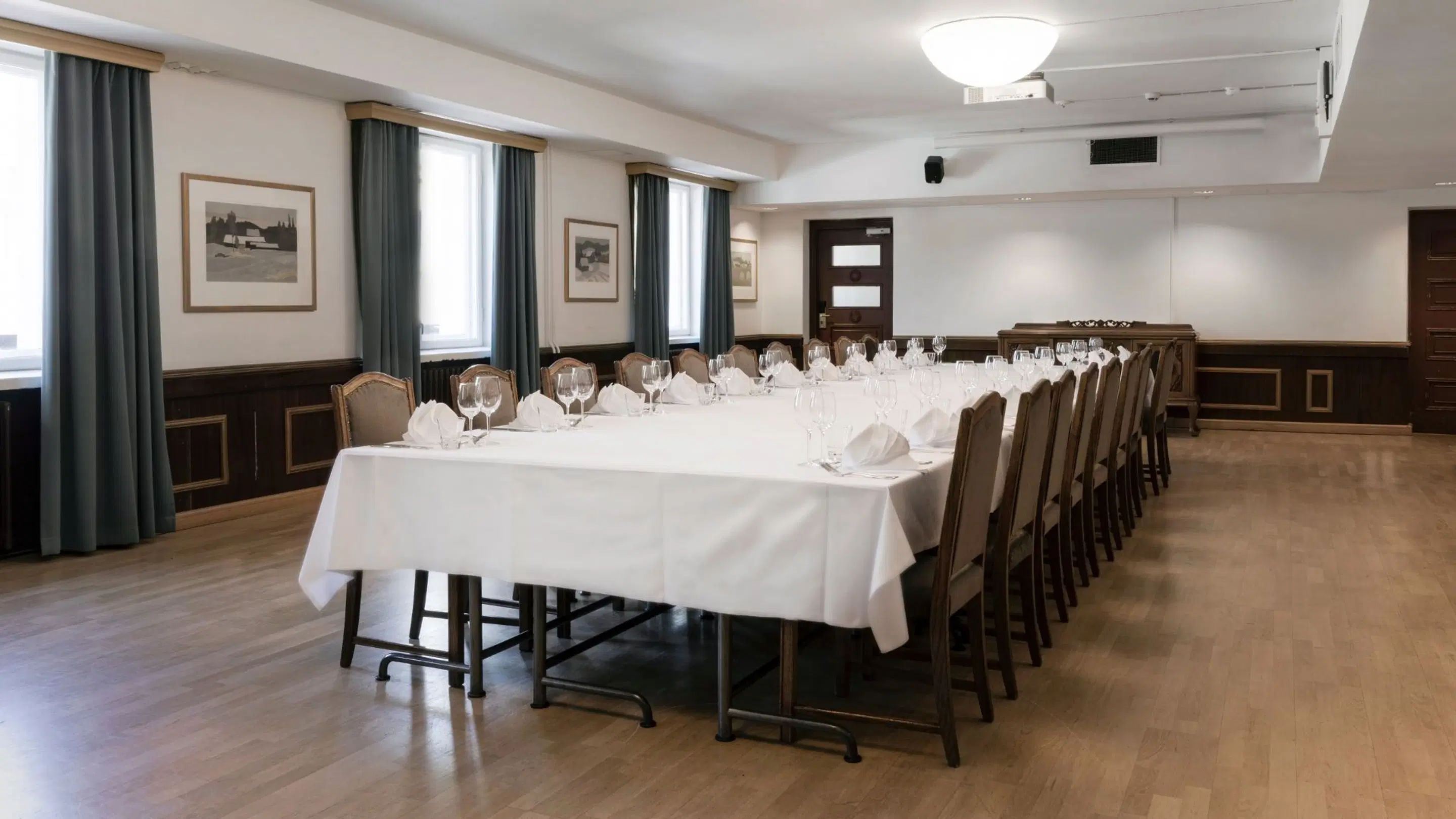 Elegant meeting room with a long table set with white tablecloth and glassware. Wooden floor, dark curtains, and even lighting throughout.