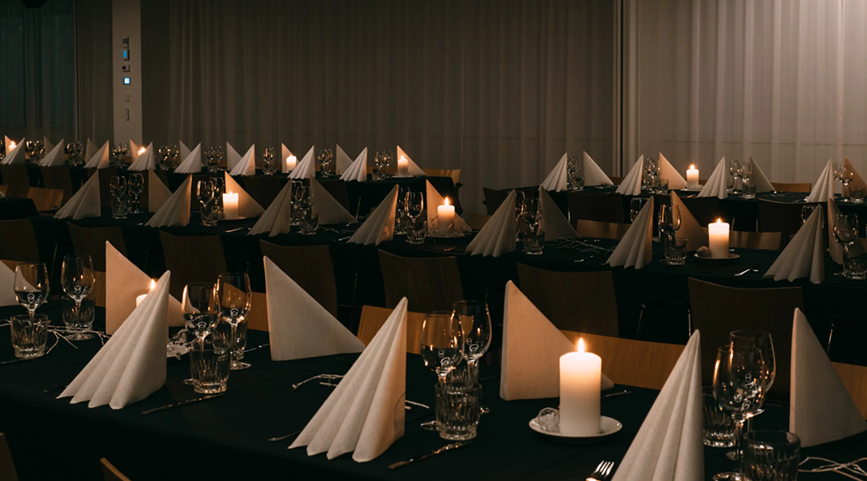 Banquet hall set with black tablecloths, folded white napkins and lit candles. Wooden chairs surround the tables.