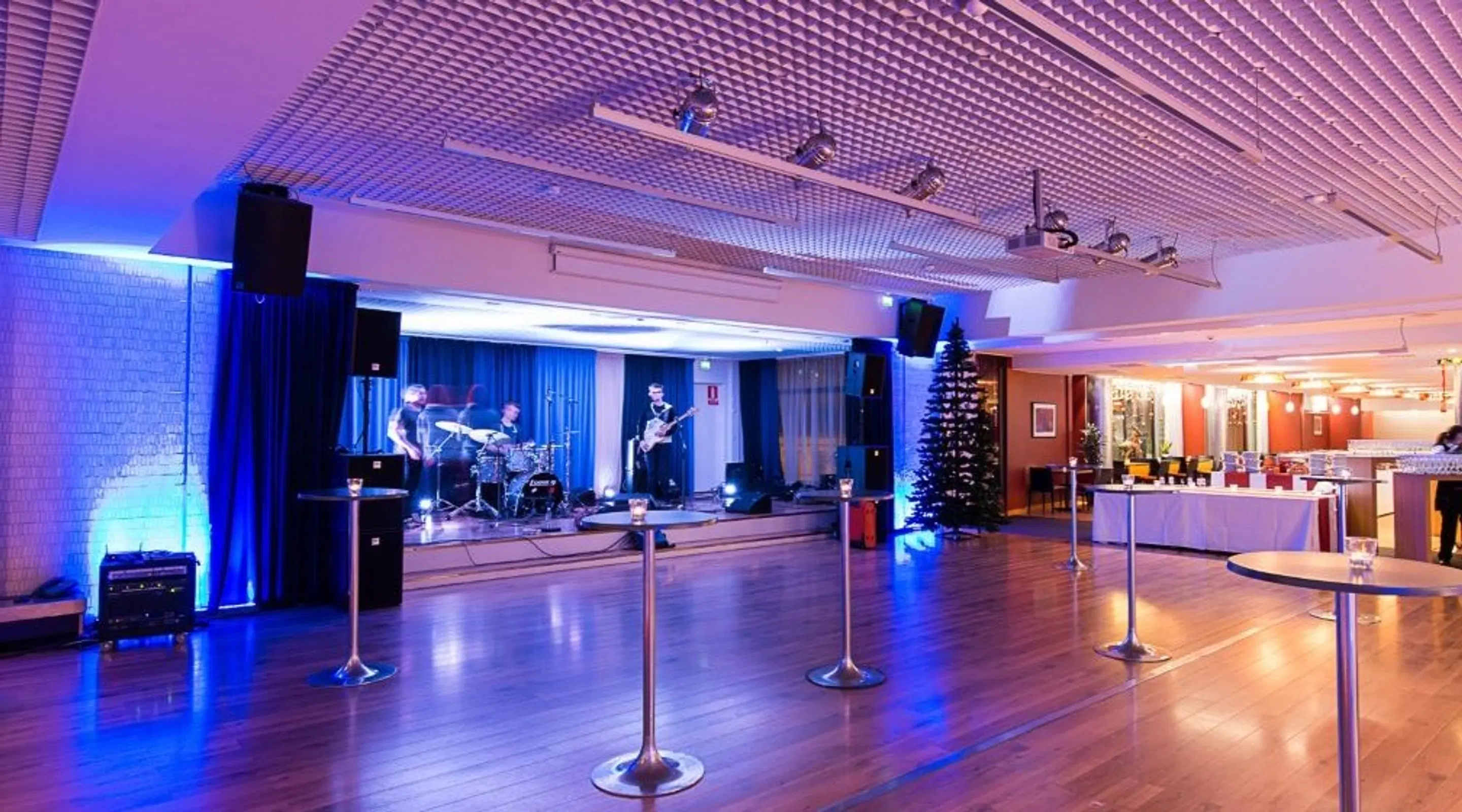 A band performing on a stage in a blue-lit hall with wooden floor, high tables, and a Christmas tree. Spacious, open area in front.