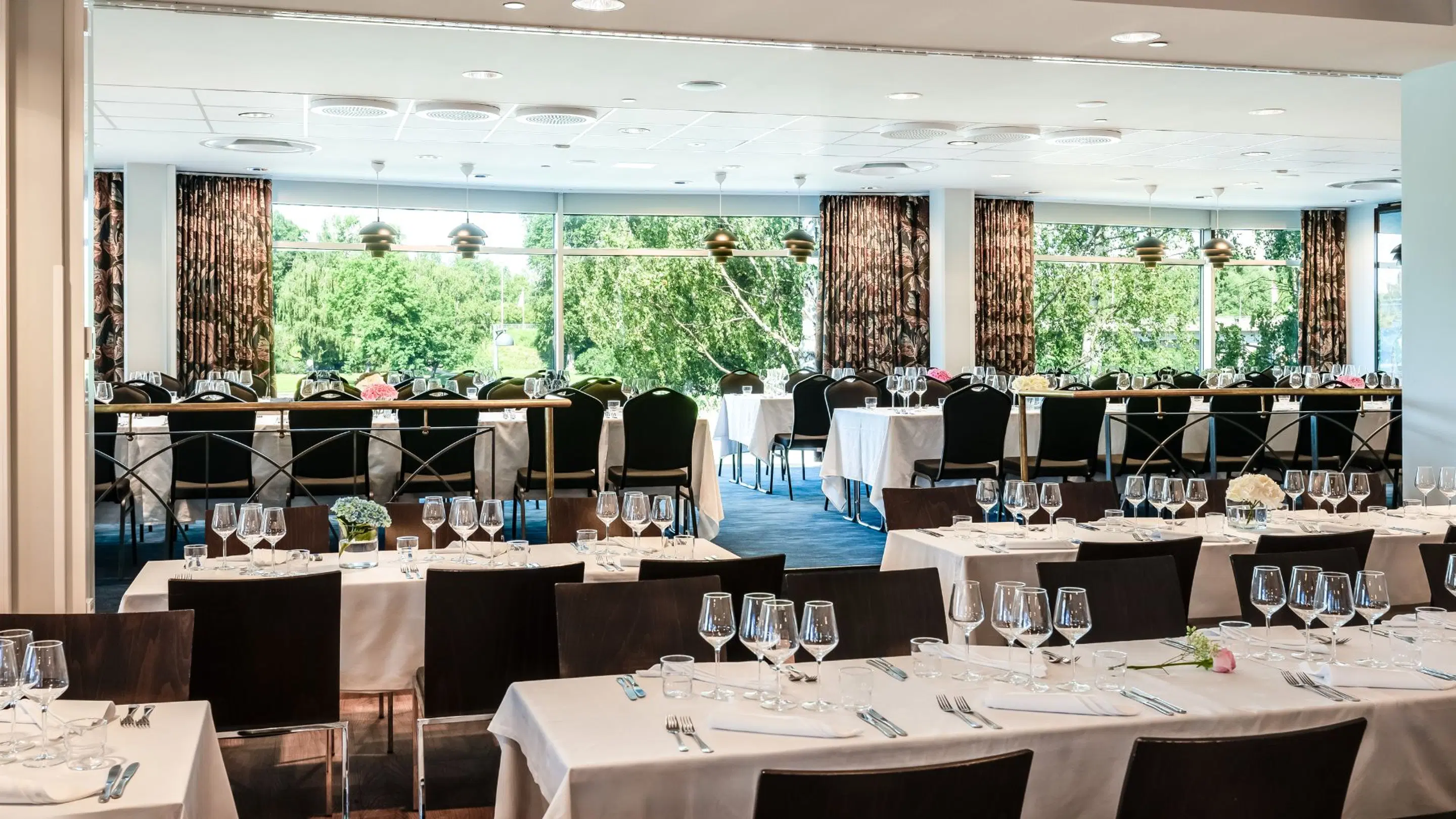 Bright dining hall with tables set for a formal event. Large windows in the background reveal a lush green forest landscape.