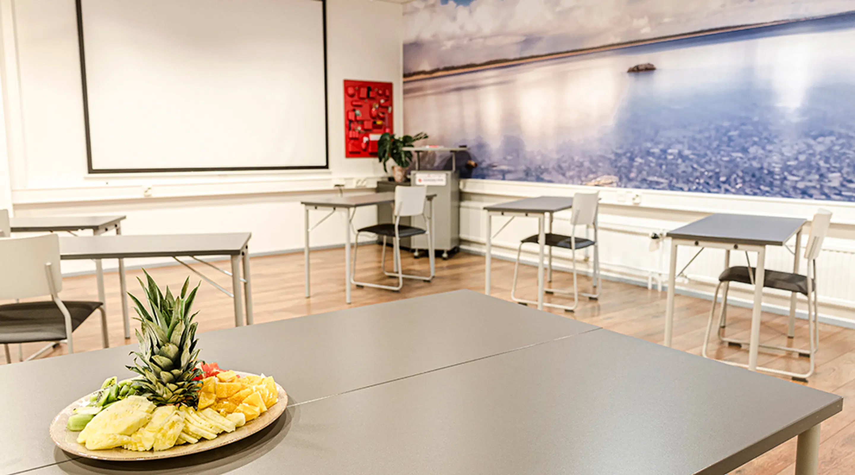 Mötesrum med separata bord, projektorduk och en fruktfat i förgrunden. Trägolv och en stor landskapsbild på väggen.